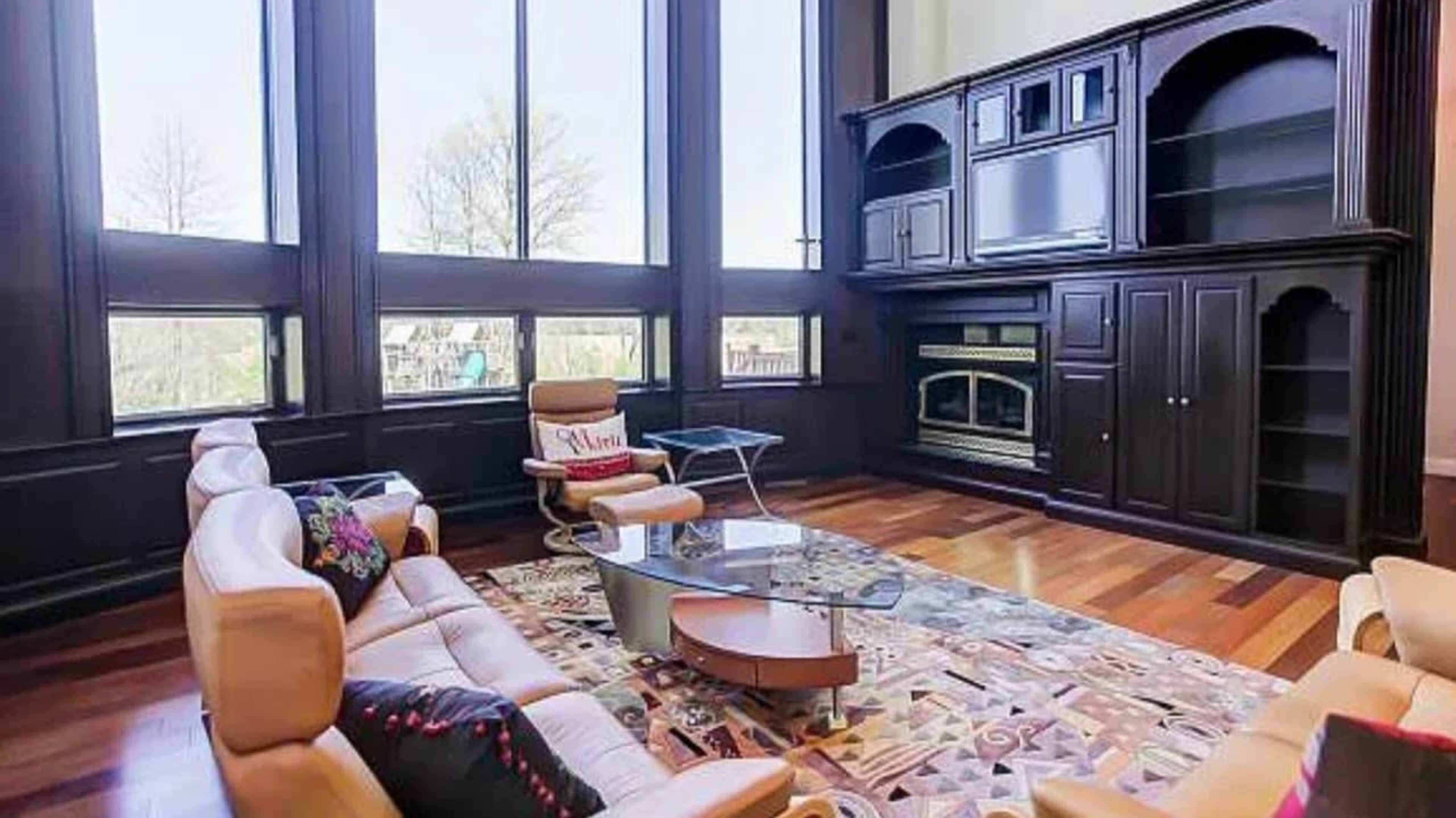 The image shows a modern living room with large windows, a cream-colored sectional sofa, a glass coffee table, and a dark wooden entertainment unit.