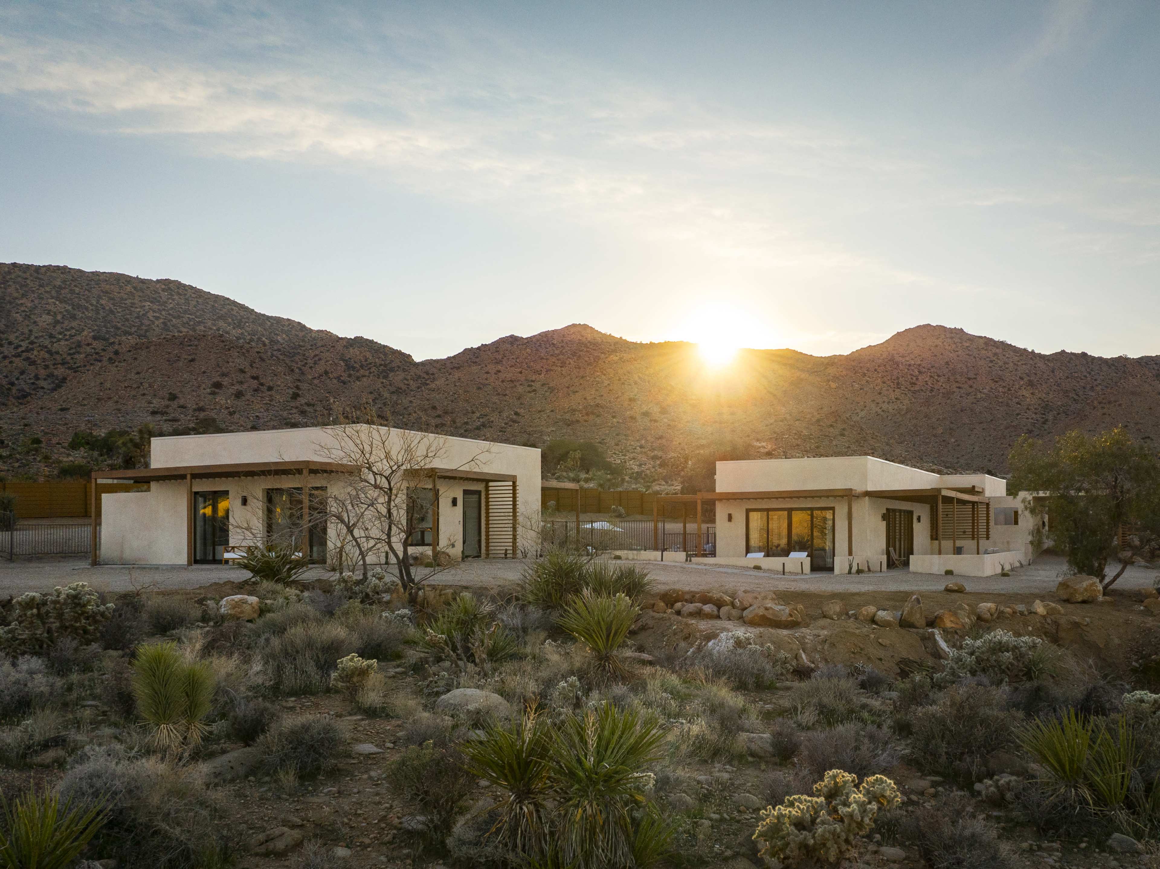 Two modern houses are positioned against a mountainous landscape at sunset, surrounded by desert vegetation.