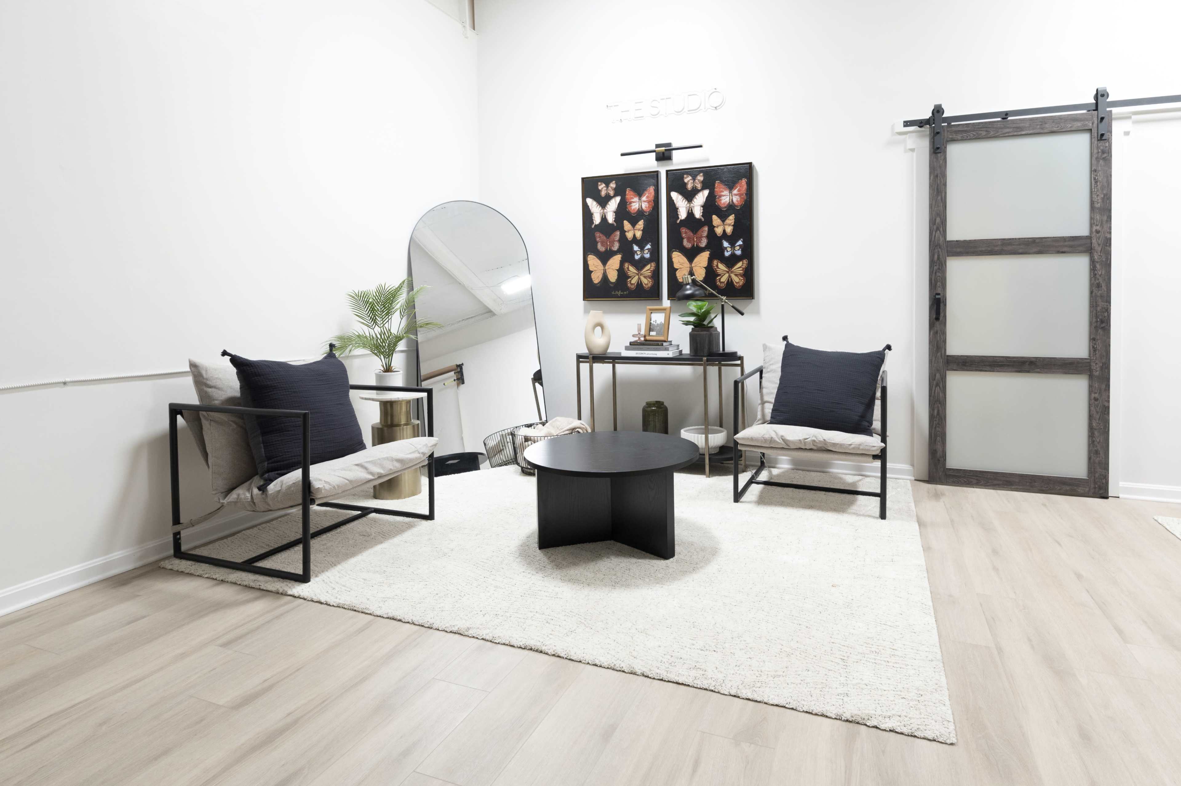 The image shows a modern living space featuring two black-framed chairs with beige cushions, a round black coffee table, a large mirror, a decorative wall with framed butterfly art, and a sliding barn door.