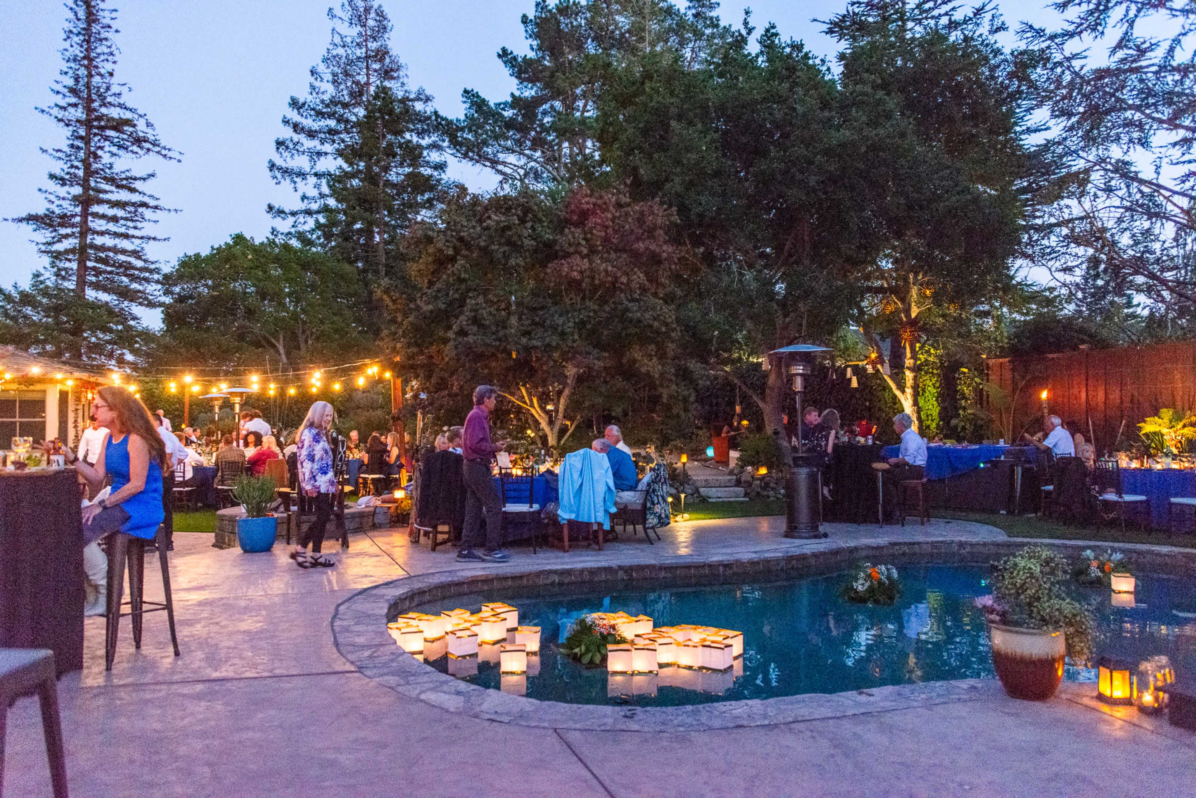 An outdoor event takes place in a garden setting with tables arranged around a pool, illuminated by string lights and floating lanterns.