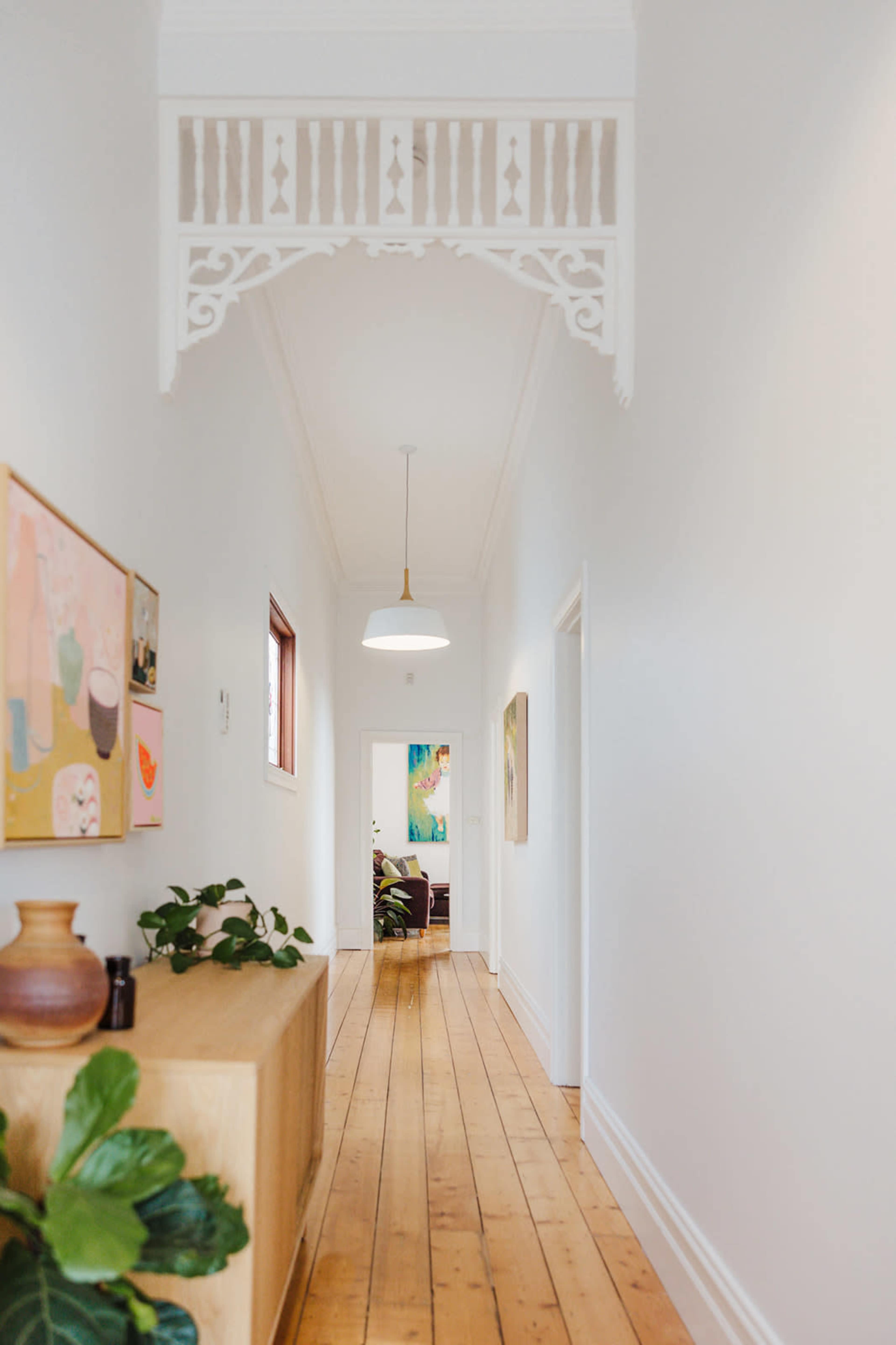 A narrow hallway features light wooden flooring, white walls, and decorative architraves, with artwork on the walls and plants on a sideboard.