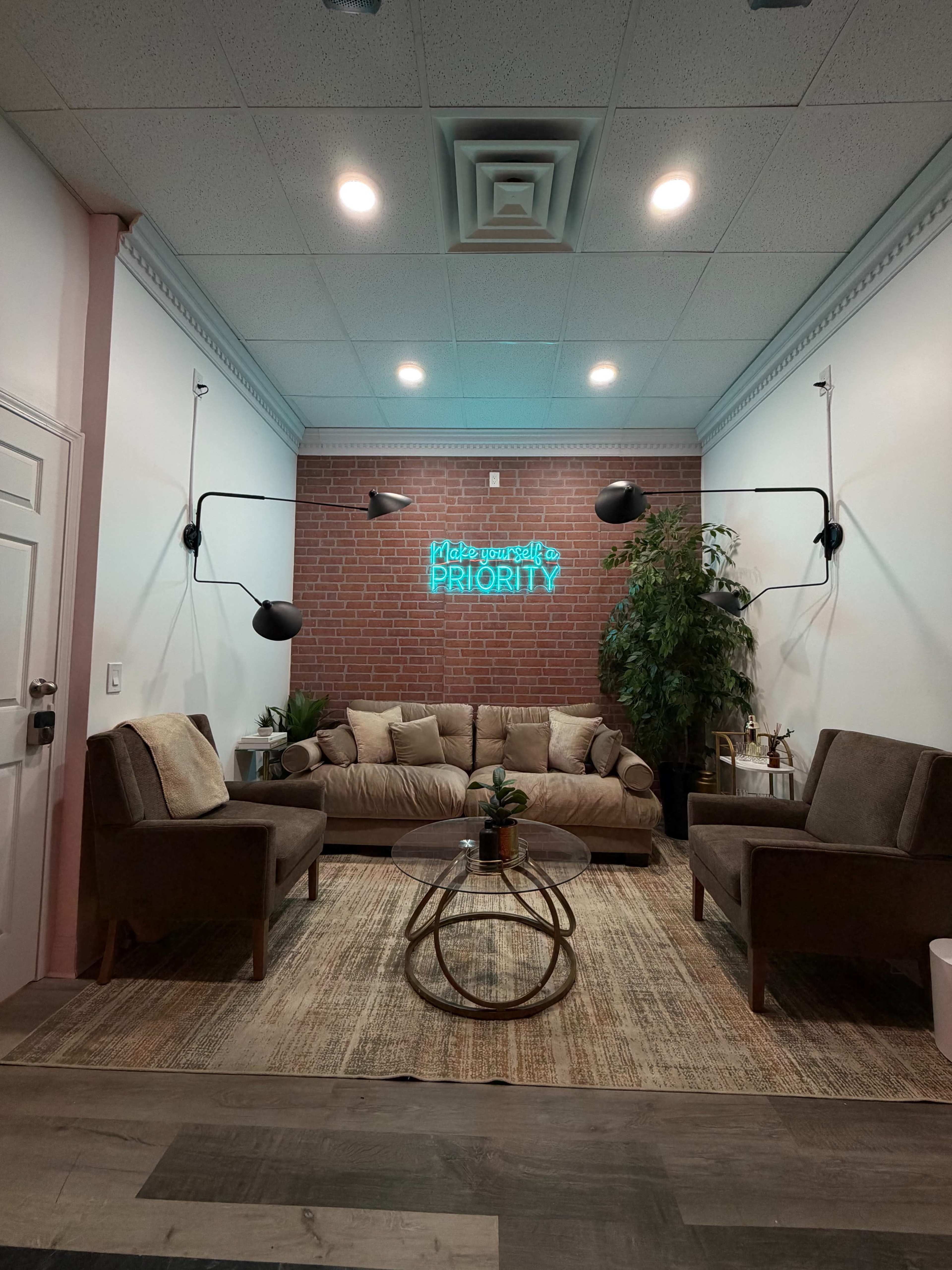 The image shows a cozy lounge area with a brick wall, a brown couch, two armchairs, a round glass coffee table, and a neon sign that reads "Make yourself a priority," beside a large green plant.