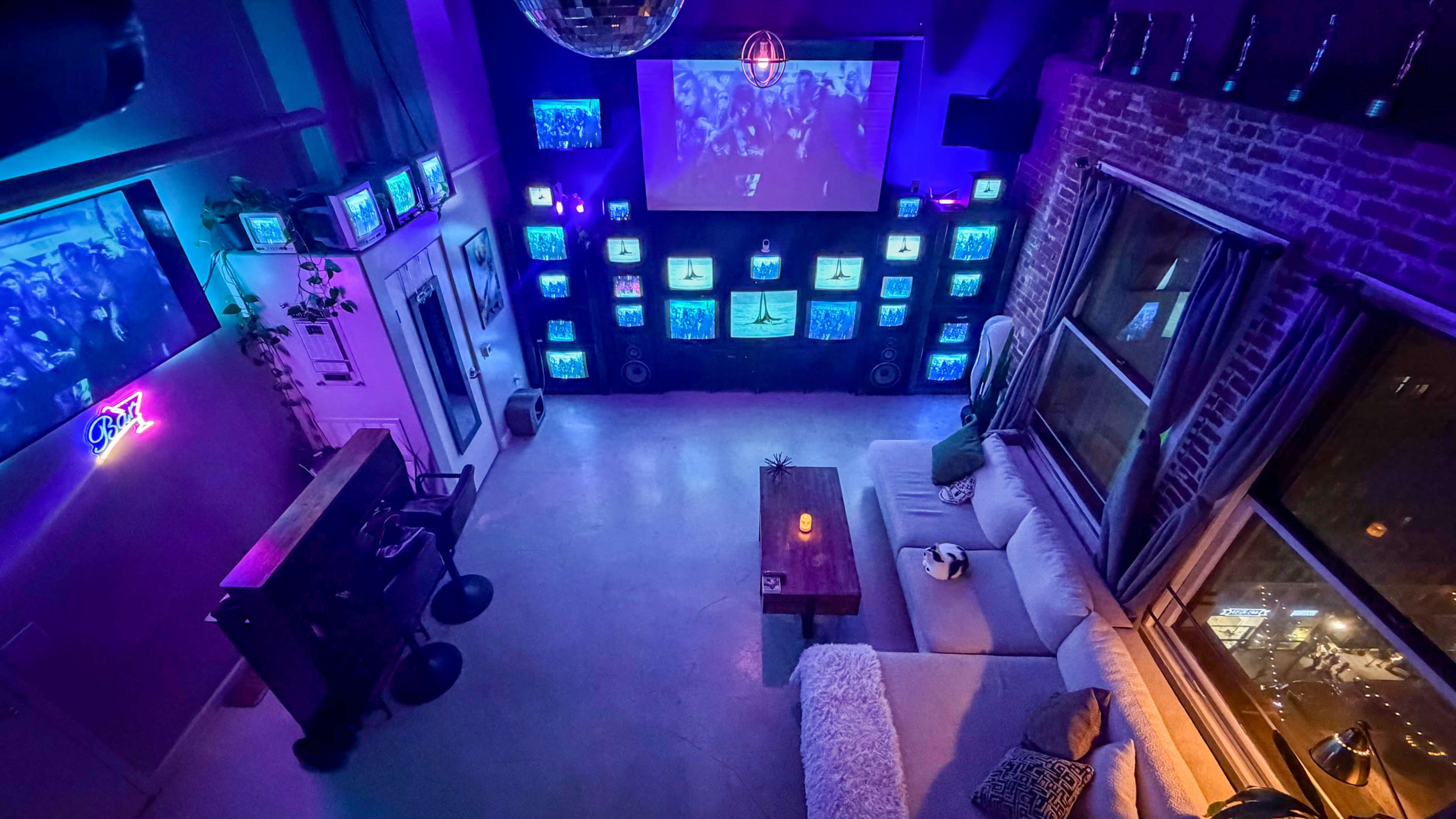 The image shows a modern living space with a large sectional sofa, a wooden coffee table with a candle, and multiple vintage television sets displaying vibrant visuals on the walls, along with a large screen projecting images in a dimly lit environment.