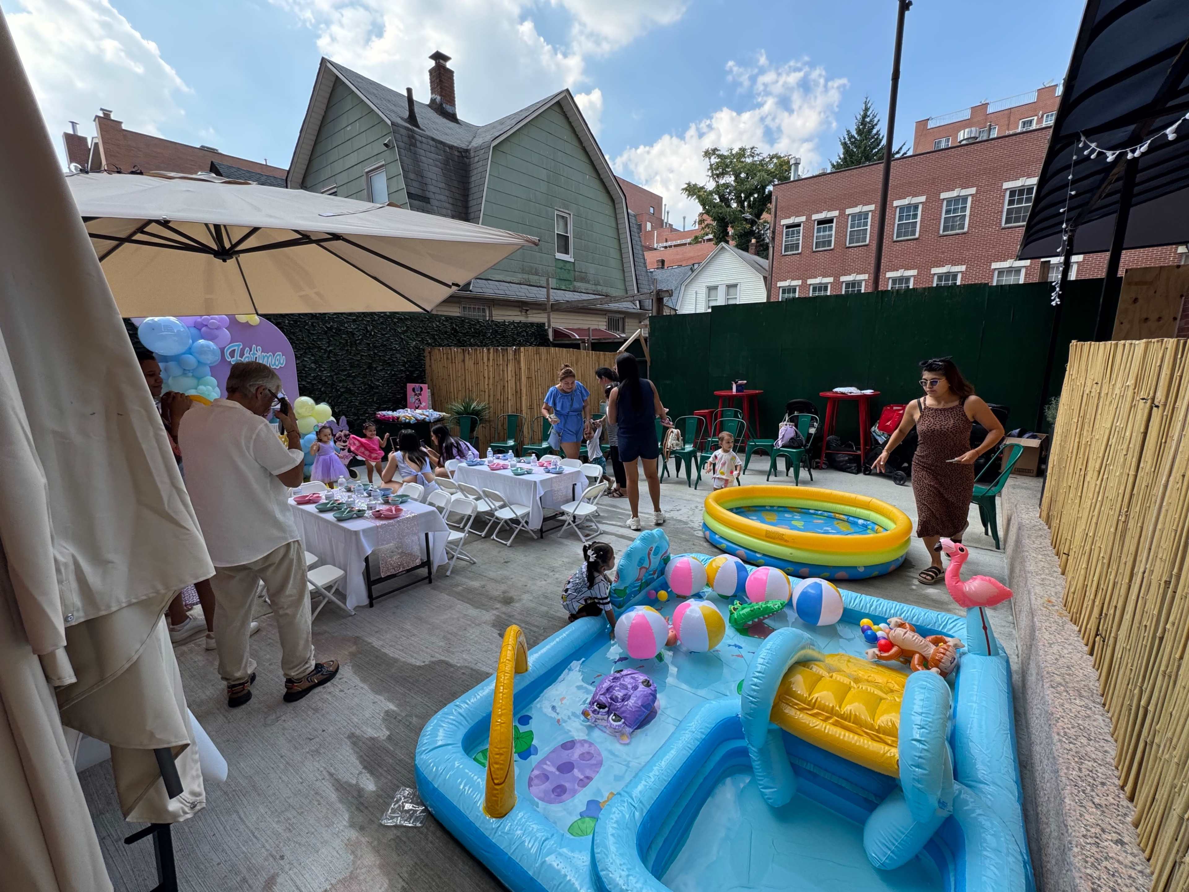 A backyard party setup features a small inflatable pool, colorful decorations, tables with chairs, and guests mingling under umbrellas.