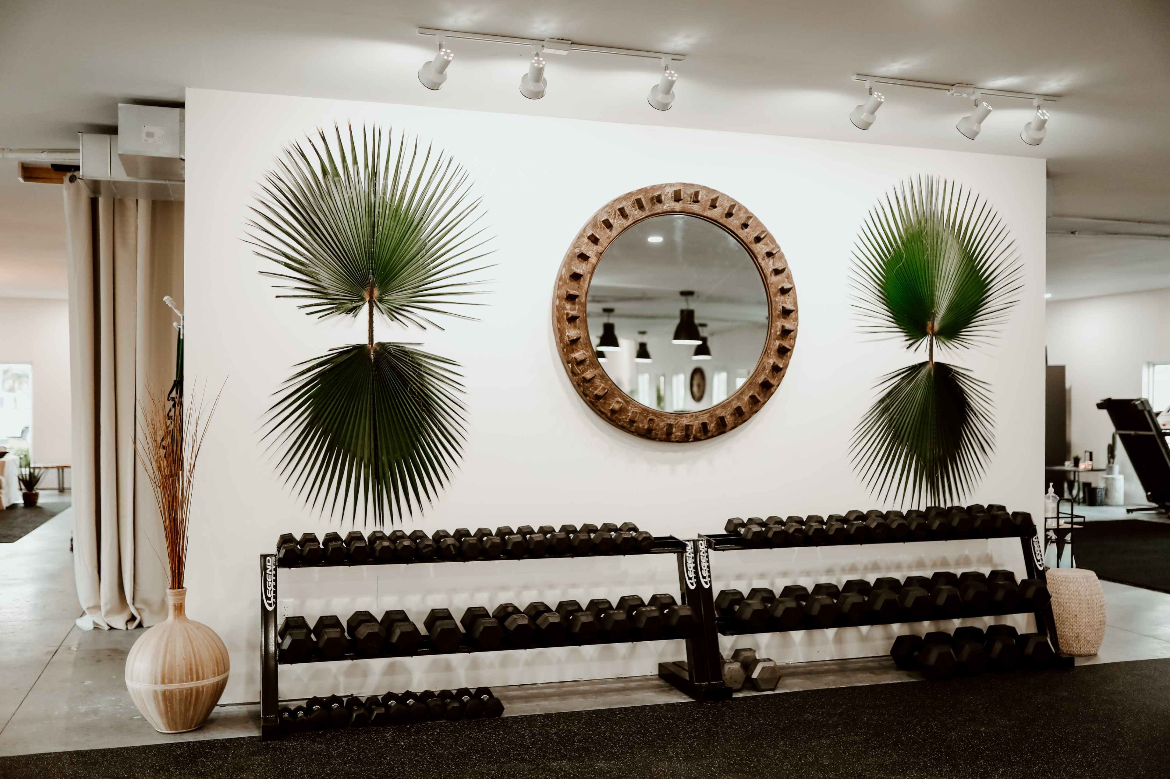 A fitness space with a wall featuring two large palm leaves on either side of a circular wooden mirror, and a rack filled with dumbbells beneath.