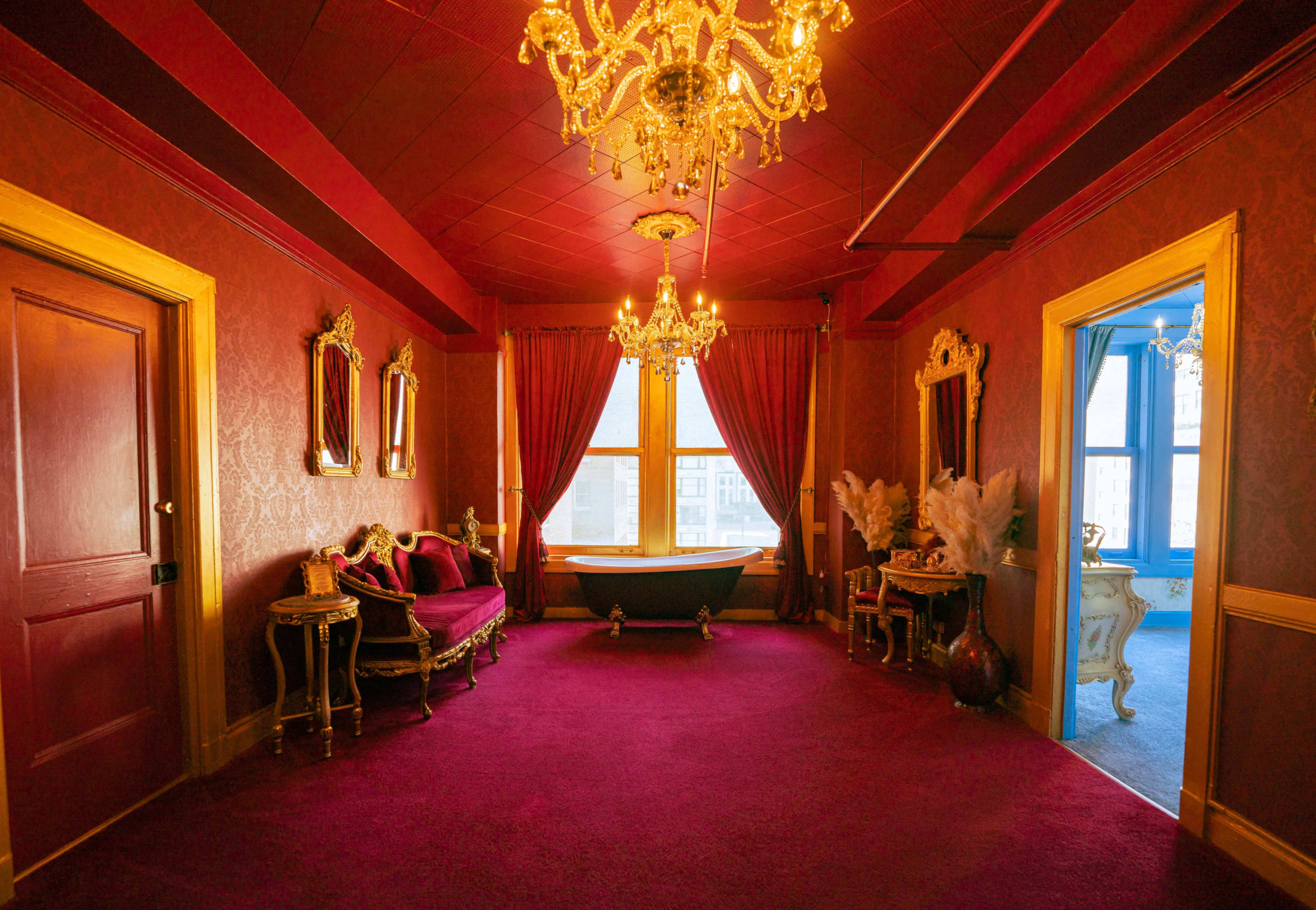 The scene shows an elegantly decorated room with red velvet walls, a plush carpet, vintage furniture, and chandeliers, featuring a large window and a bathtub.