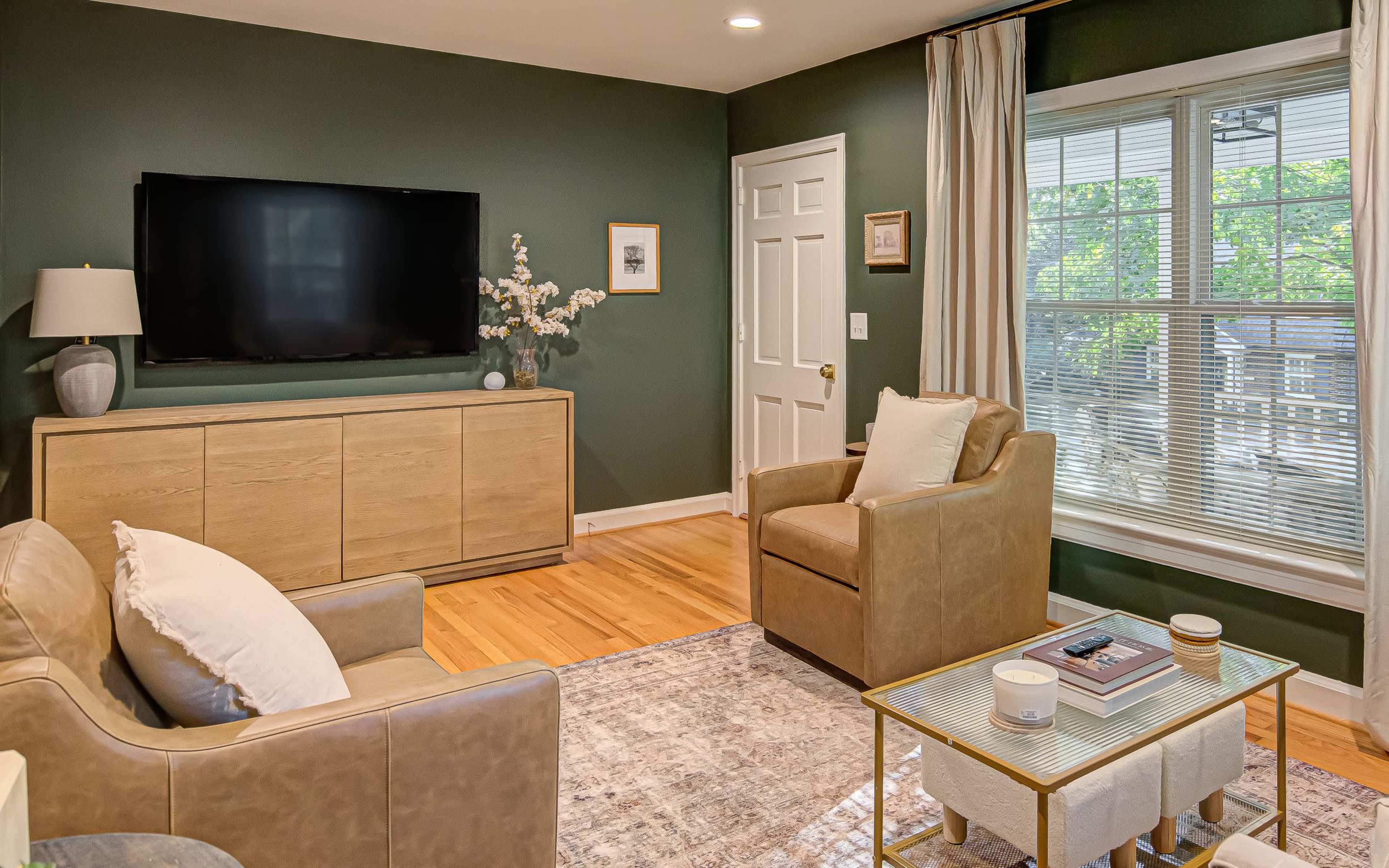A modern living room features a wall-mounted television, a light wood cabinet, two armchairs, and a coffee table, all set against a dark green wall.