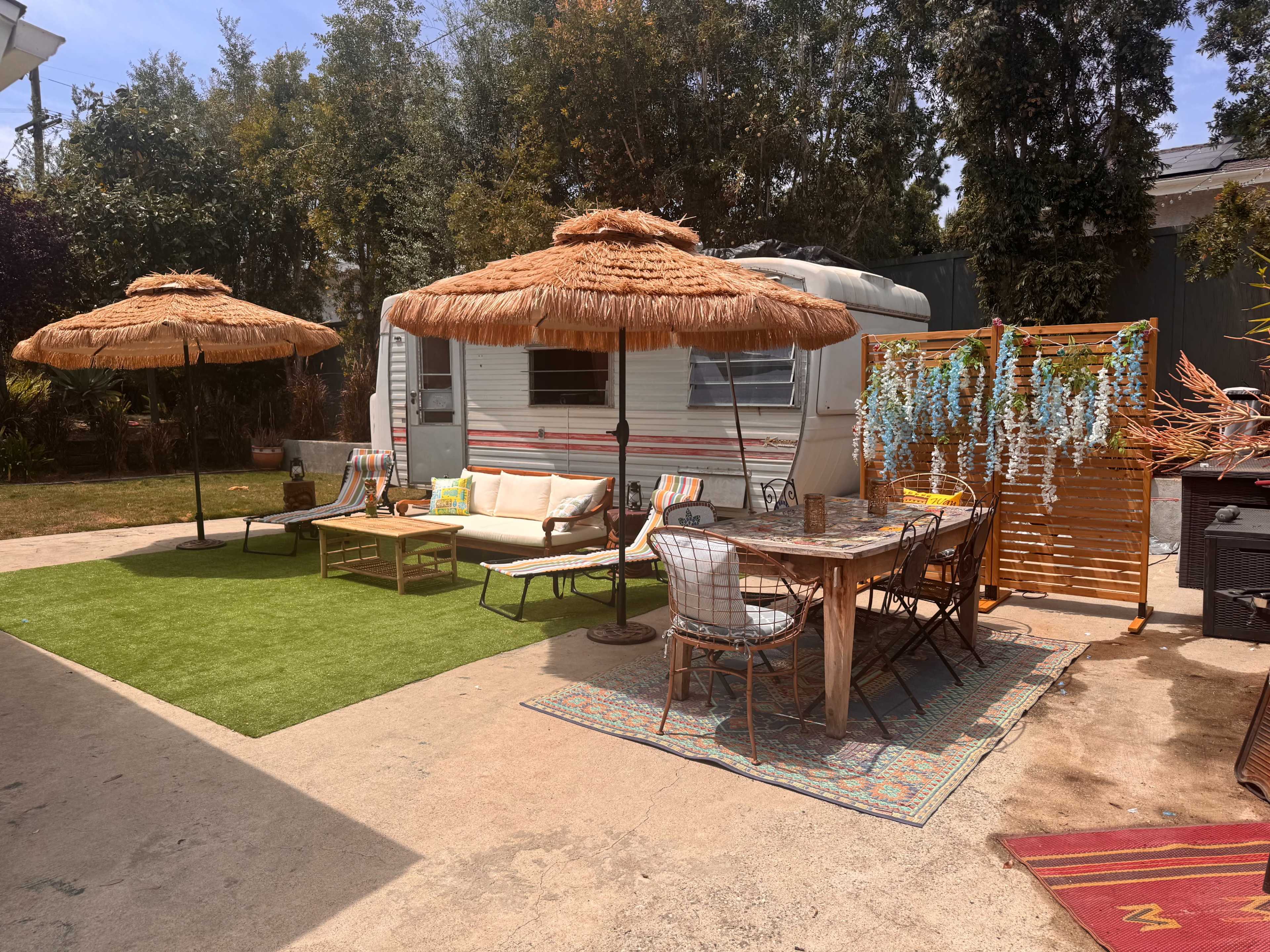 A cozy outdoor space features a camper trailer, two thatched umbrellas, a seating area with a couch, a dining table with chairs, and decorative greenery.