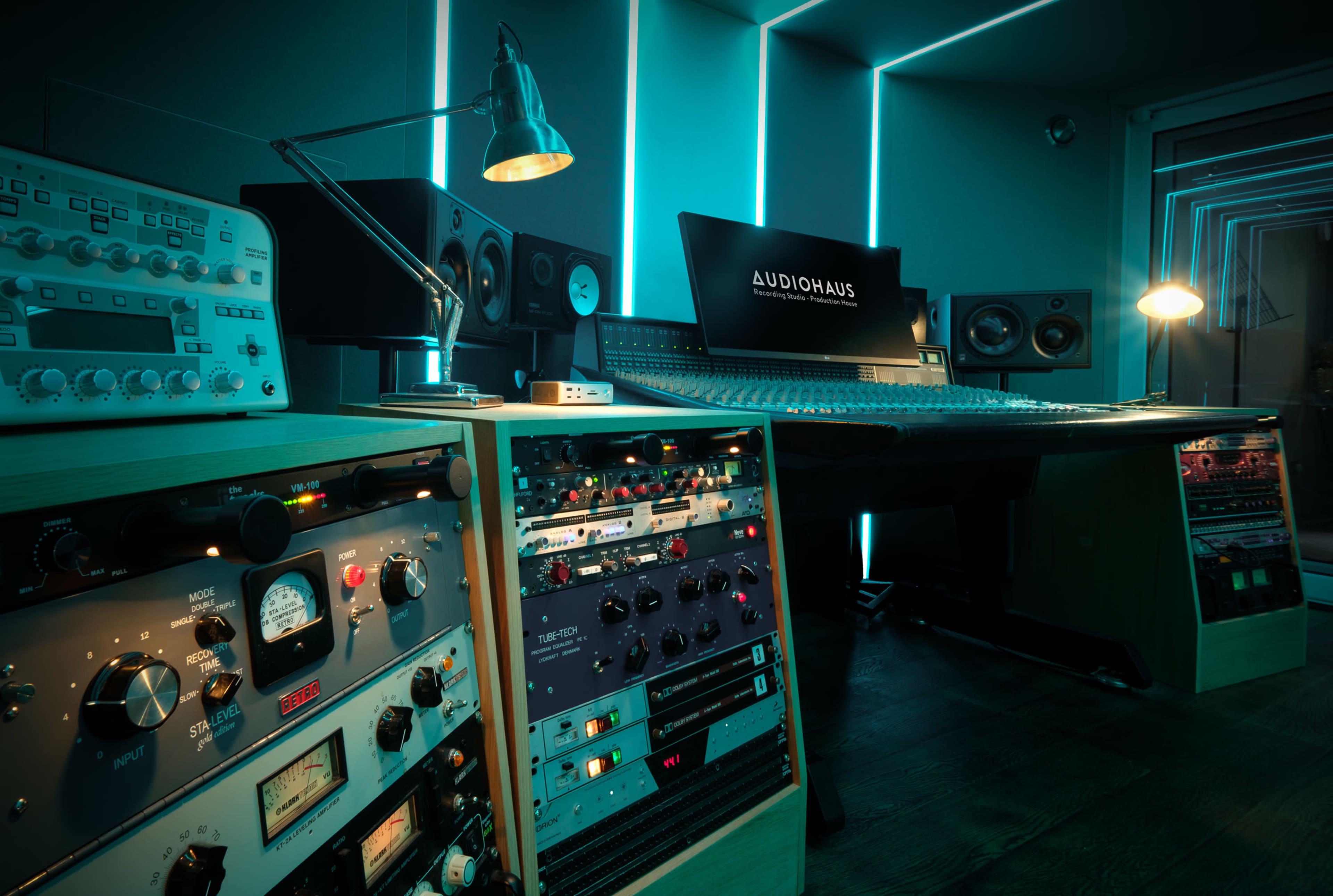 The image shows a modern audio recording studio with a central mixing console, rack-mounted equipment, and ambient lighting.