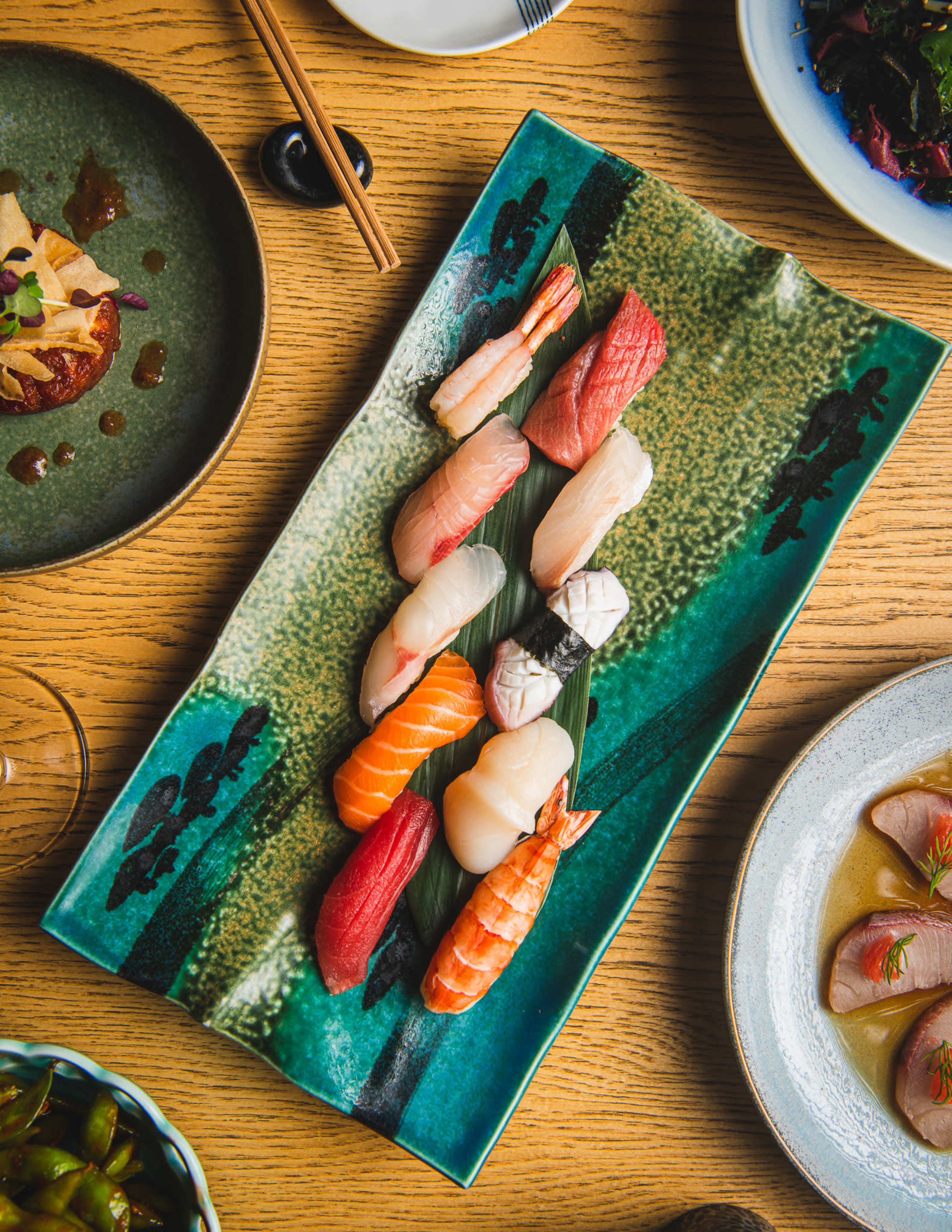 A colorful selection of sushi and sashimi is presented on a decorative green plate, surrounded by other dishes on a wooden table.