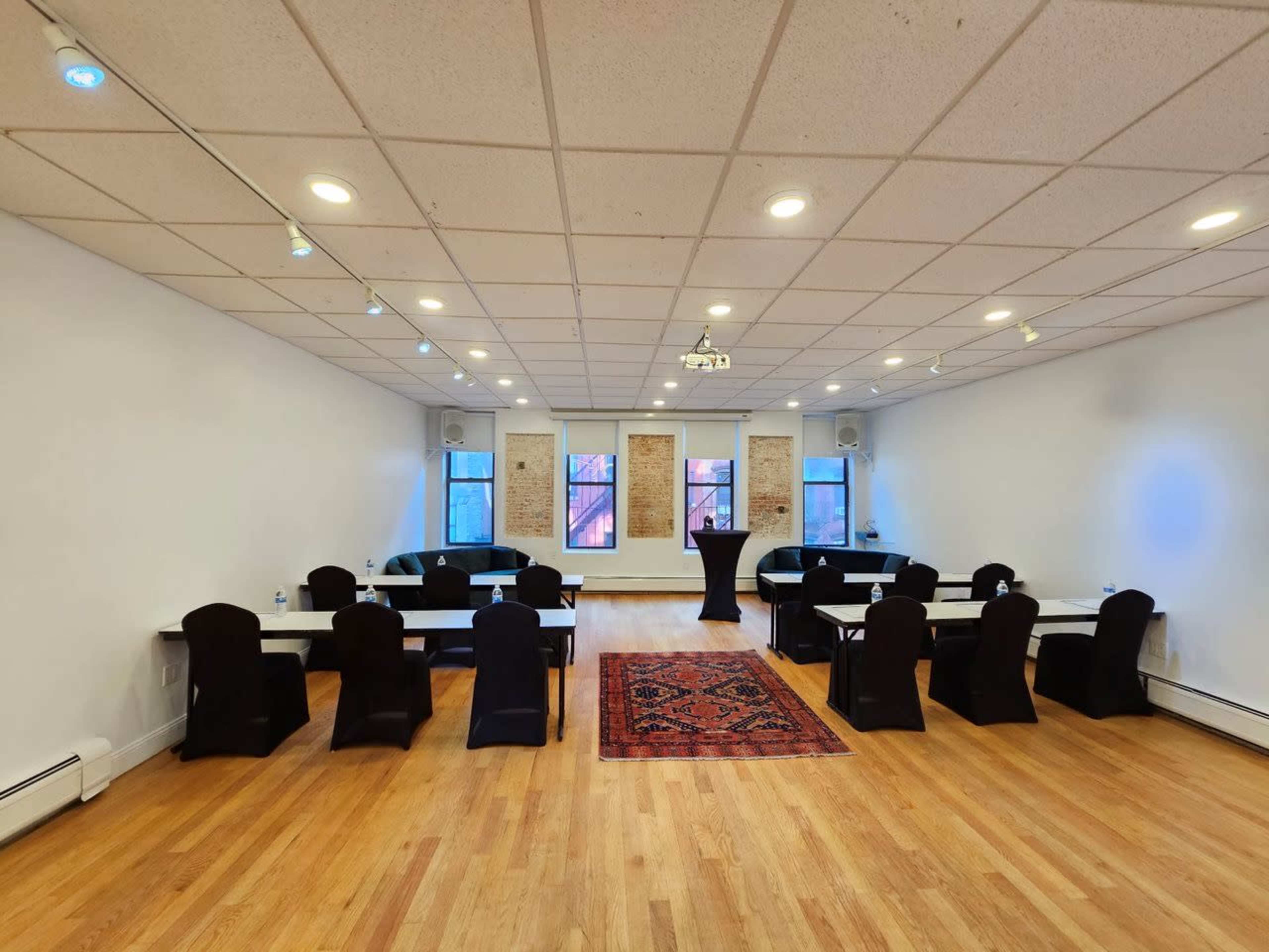 A spacious meeting room with tables arranged in clusters, black chairs, and an area rug, featuring windows on one wall that allow natural light.