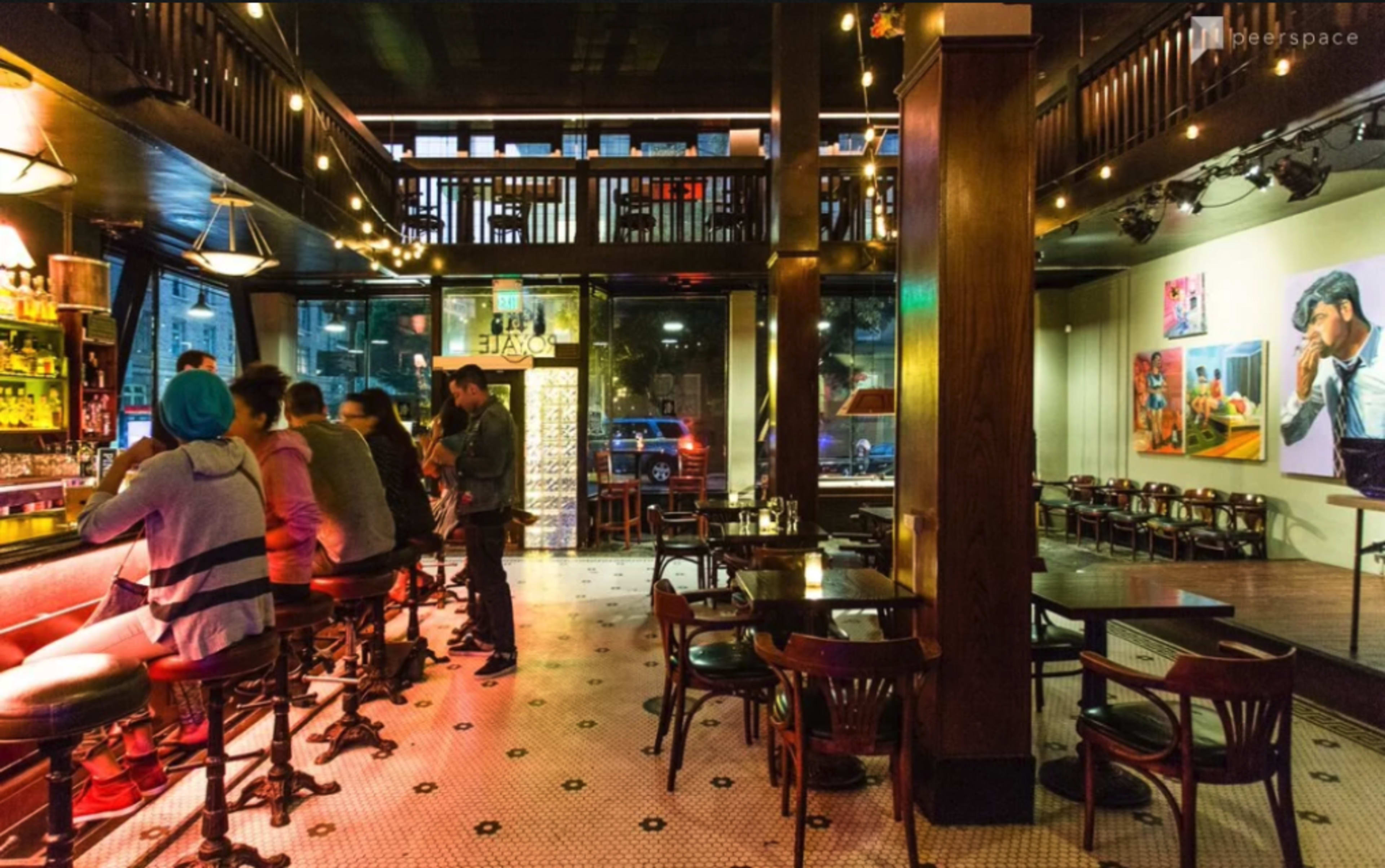 A bustling bar interior with patrons seated at the counter and tables, illuminated by warm lighting and decorated with artwork on the walls.