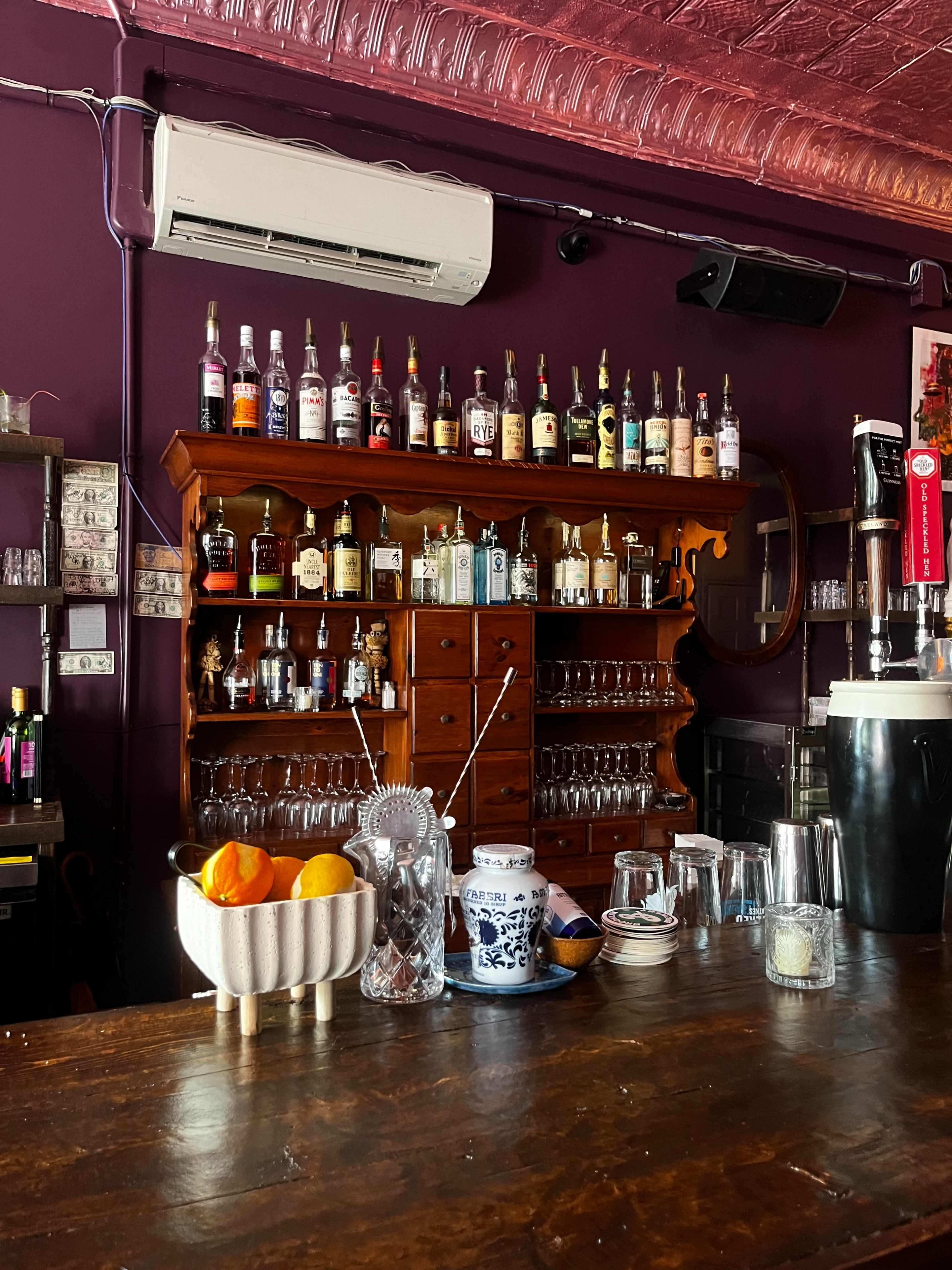 A bar with a wooden shelf displaying various bottles of alcohol and a counter featuring a bowl of oranges and glassware.
