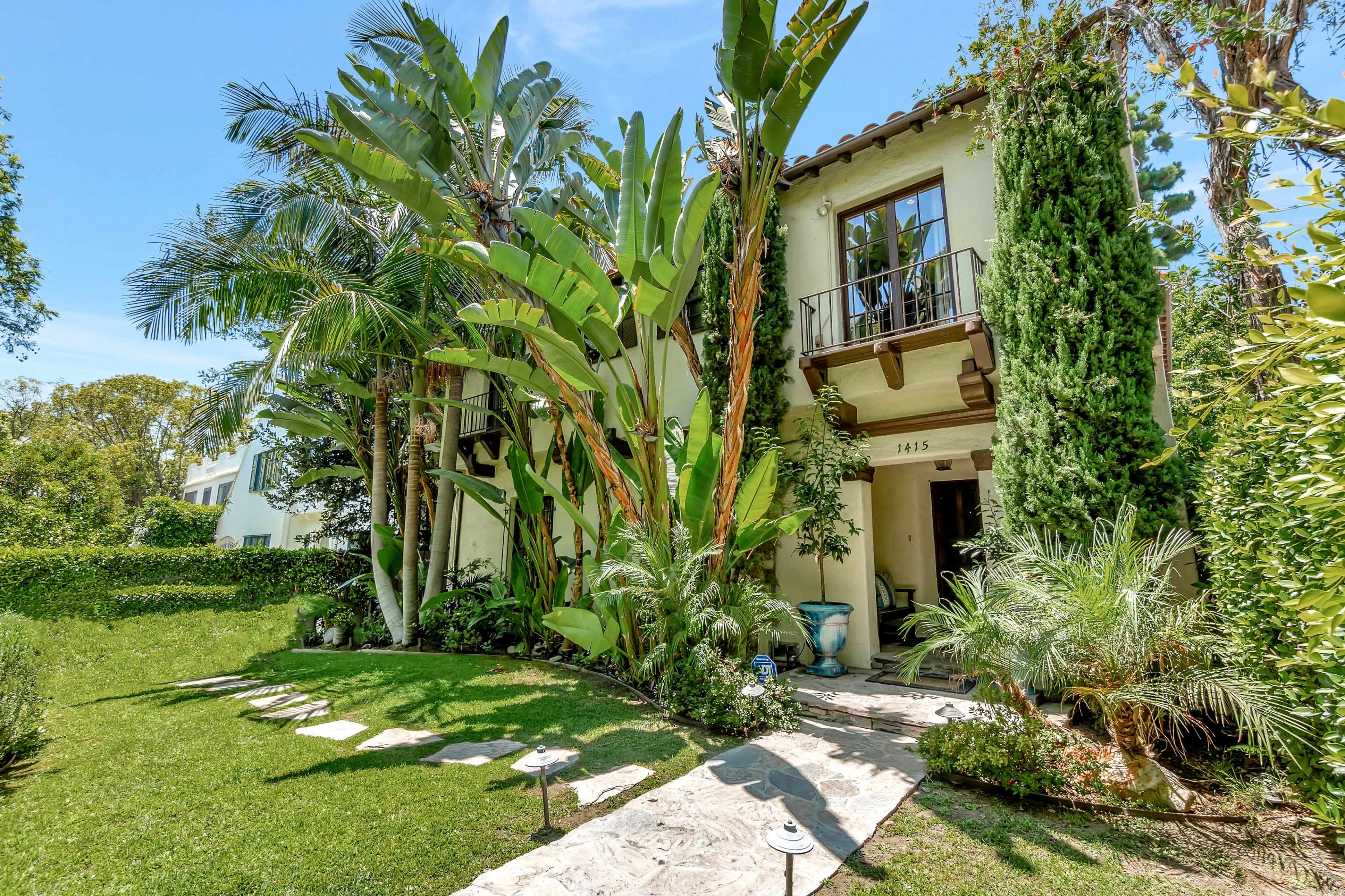 The image shows a two-story house surrounded by lush greenery and tropical plants, with a stone pathway leading to the entrance.