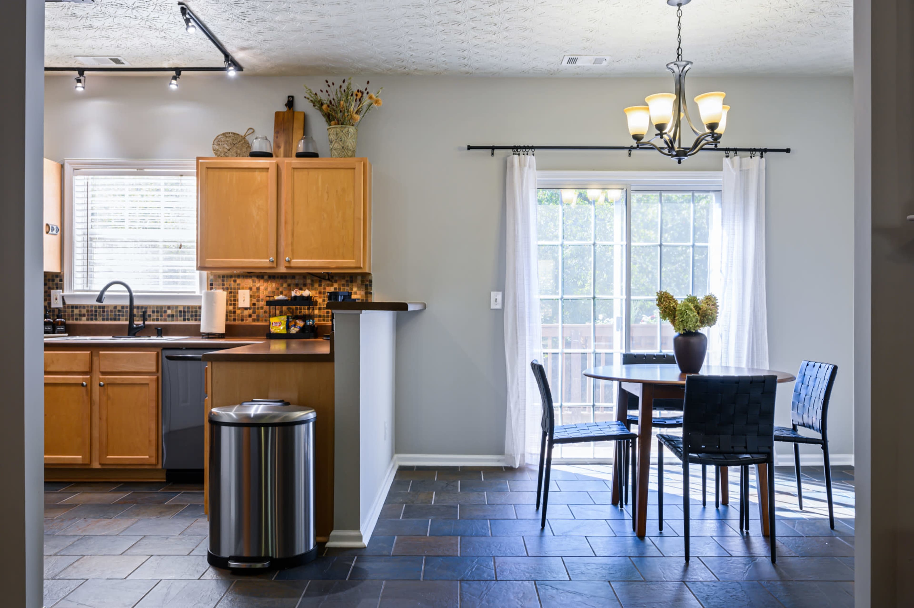 A modern kitchen and dining area with wooden cabinets, a round table set for four, and a large window leading to a patio.
