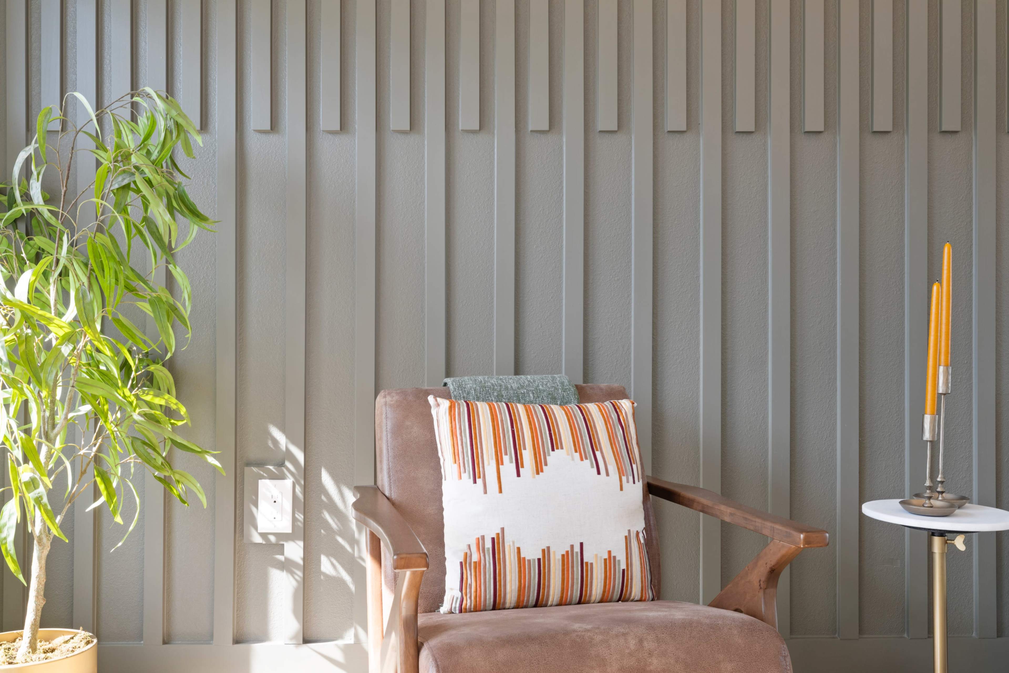 A brown armchair with a striped pillow sits beside a small white table and a potted plant against a patterned gray wall.
