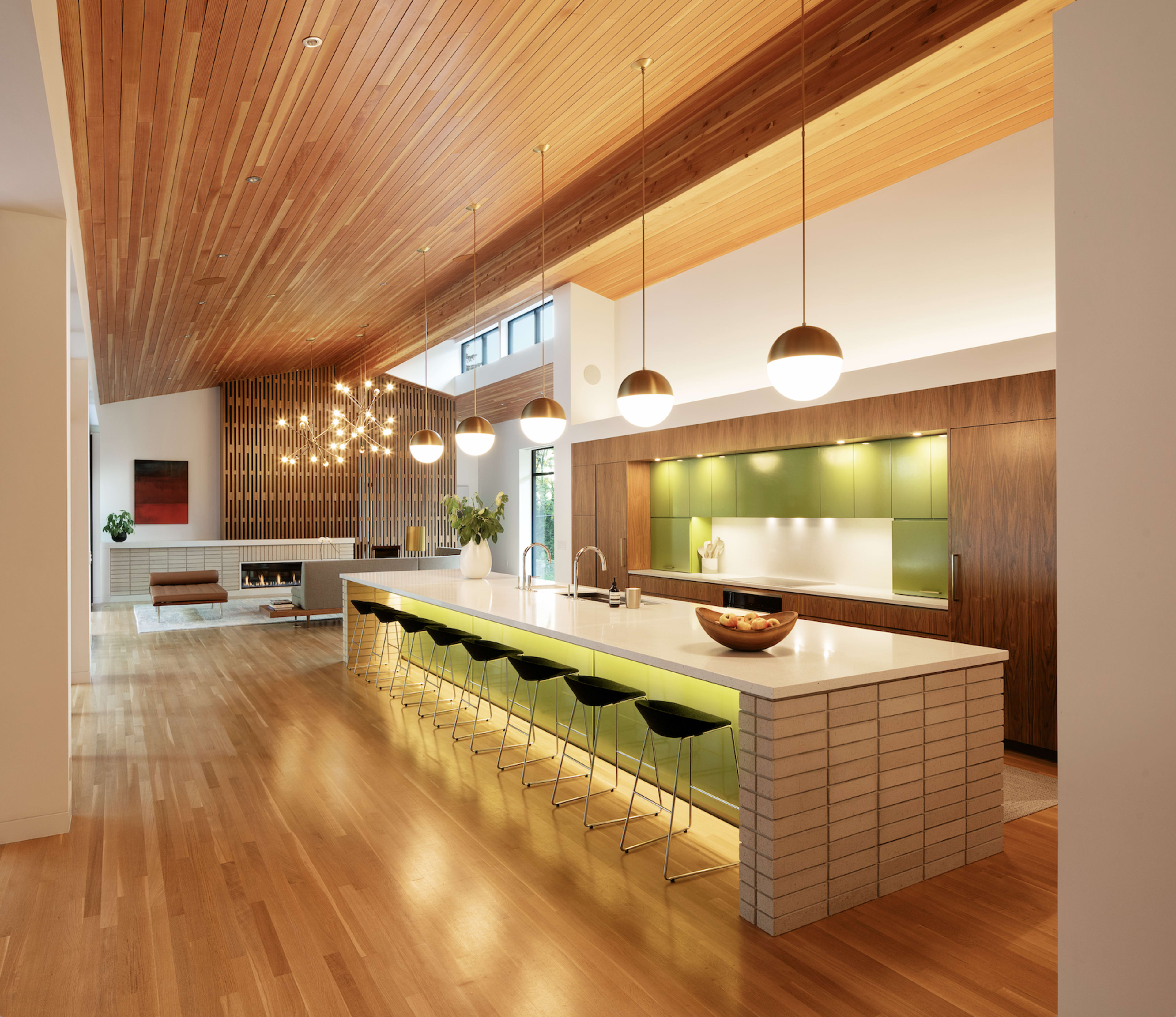 A spacious modern kitchen features a large island with green stools, pendant lights, and wood paneling overhead.