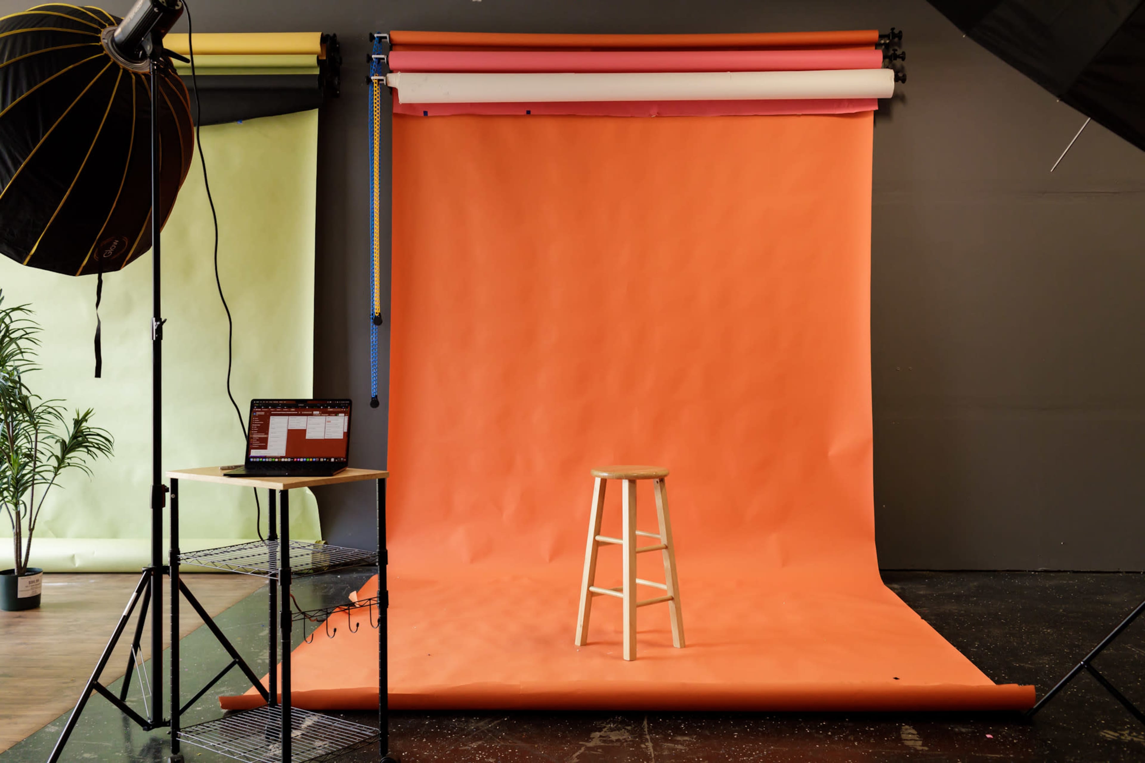 A wooden stool sits in front of a vibrant orange backdrop in a photography studio with additional colorful backdrops visible.