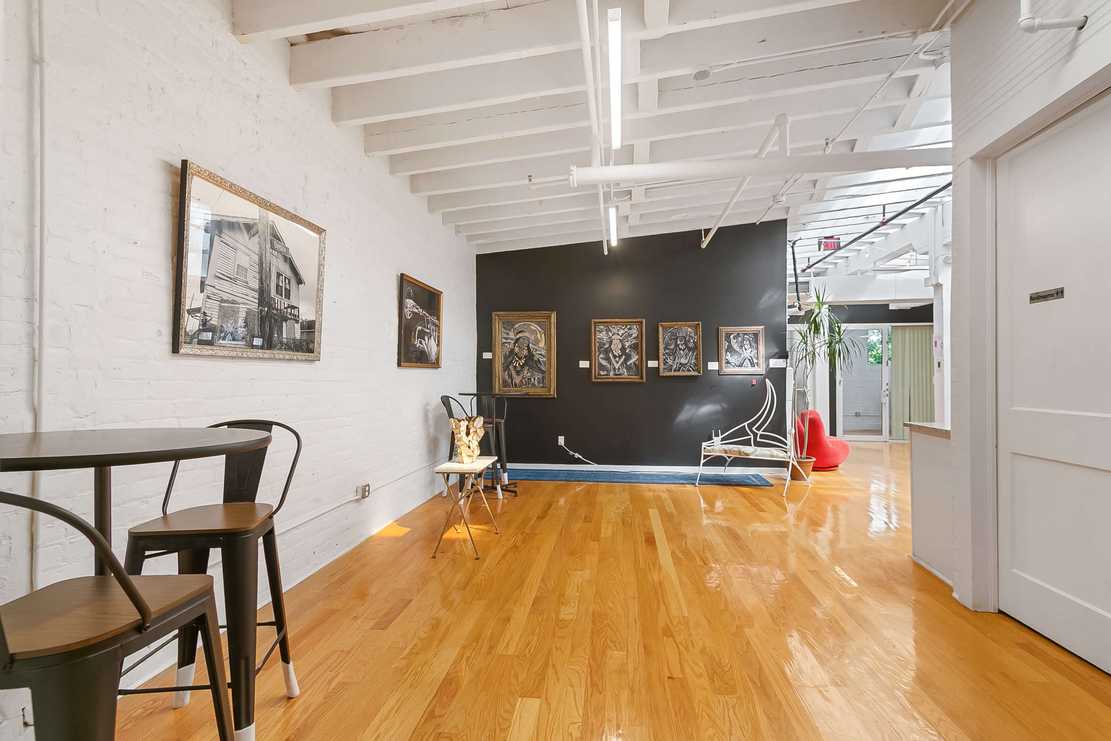 The image shows a spacious room with hardwood flooring, featuring a mix of black and white walls, contemporary art on display, and a seating area with modern furniture.