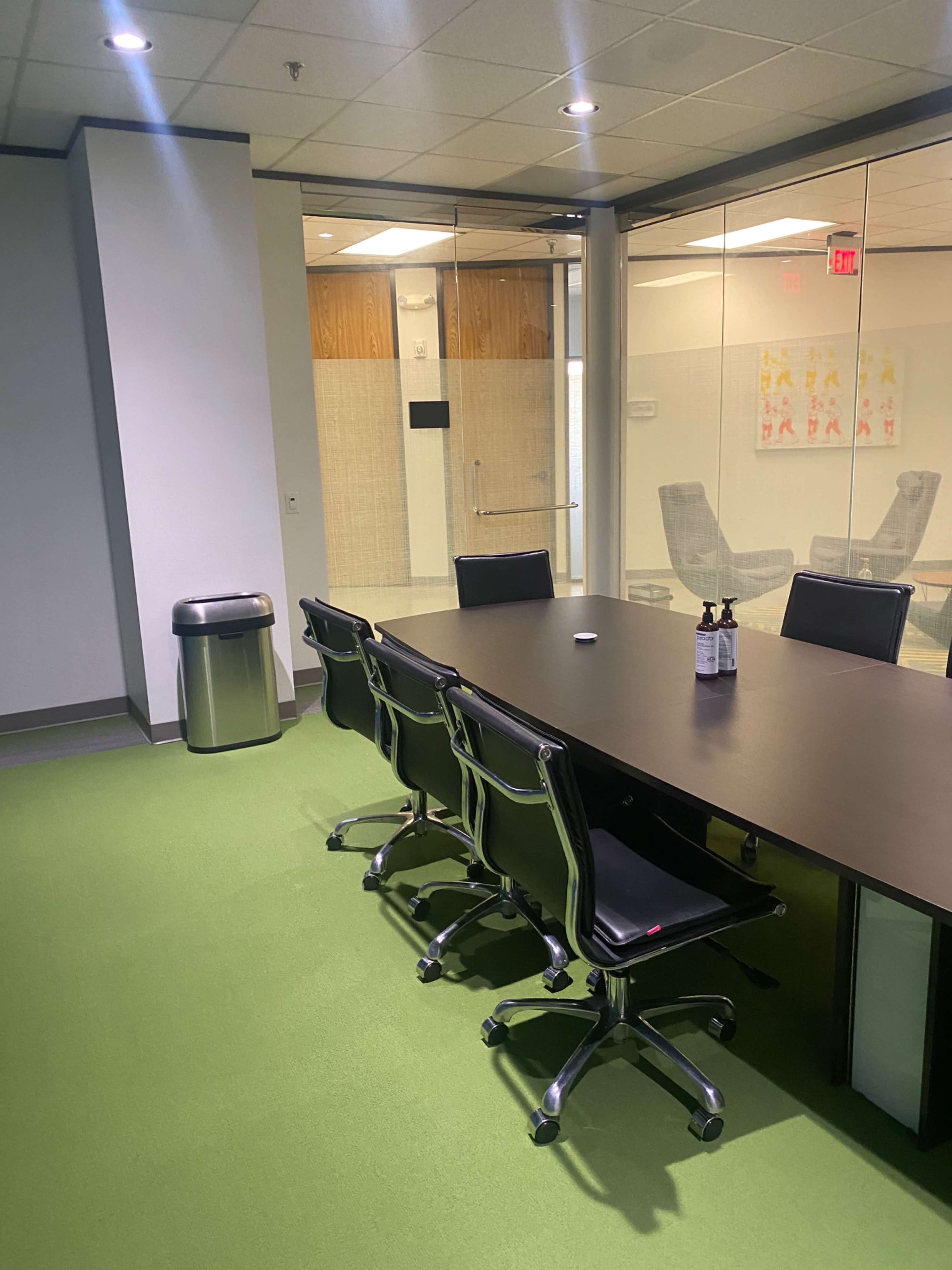 A conference room featuring a large, dark table surrounded by black rolling chairs, with green carpet and a glass-walled meeting space visible in the background.