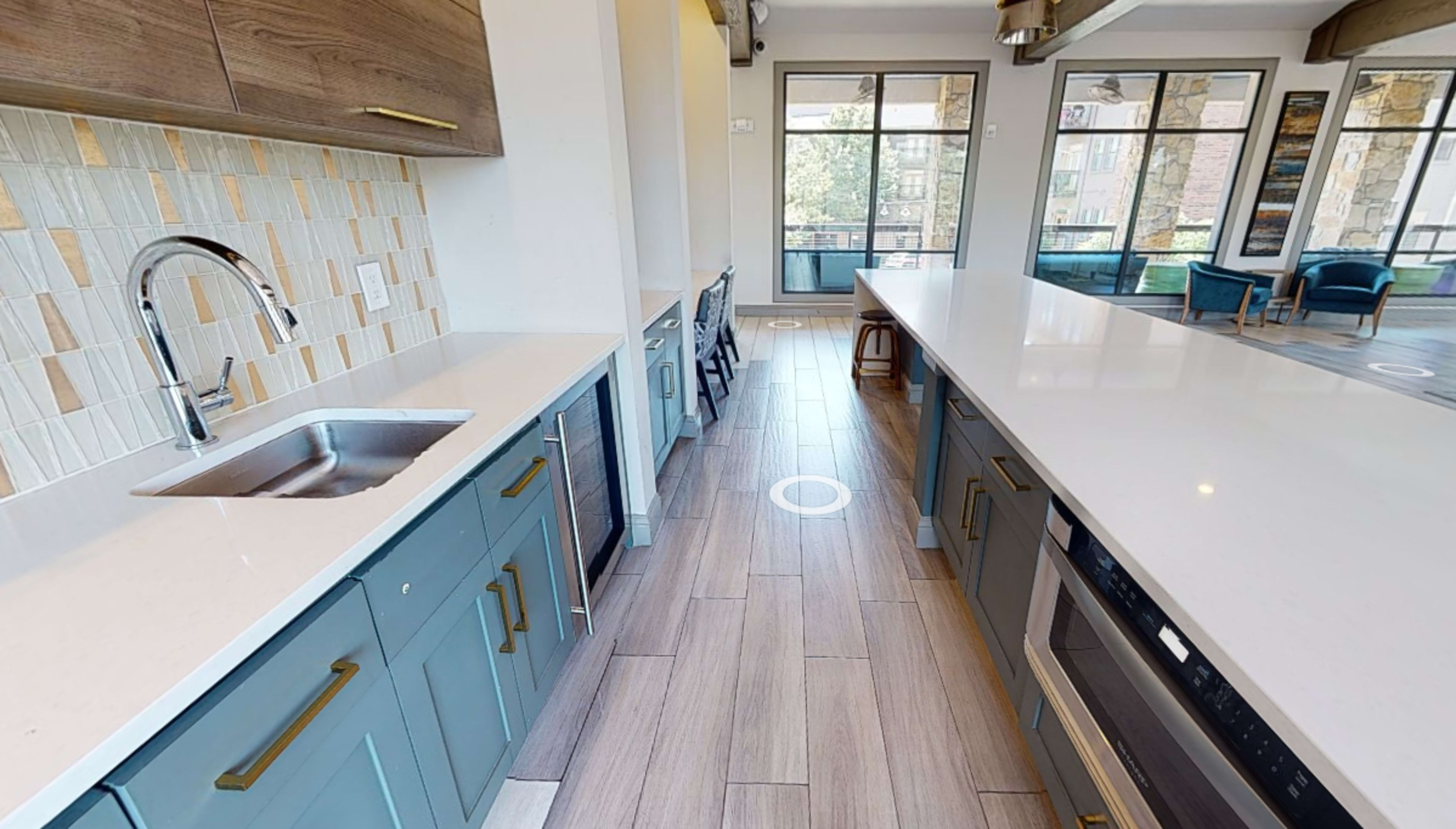 A modern kitchen with blue cabinetry, a double sink, and a large island overlooking a bright living area through large windows.