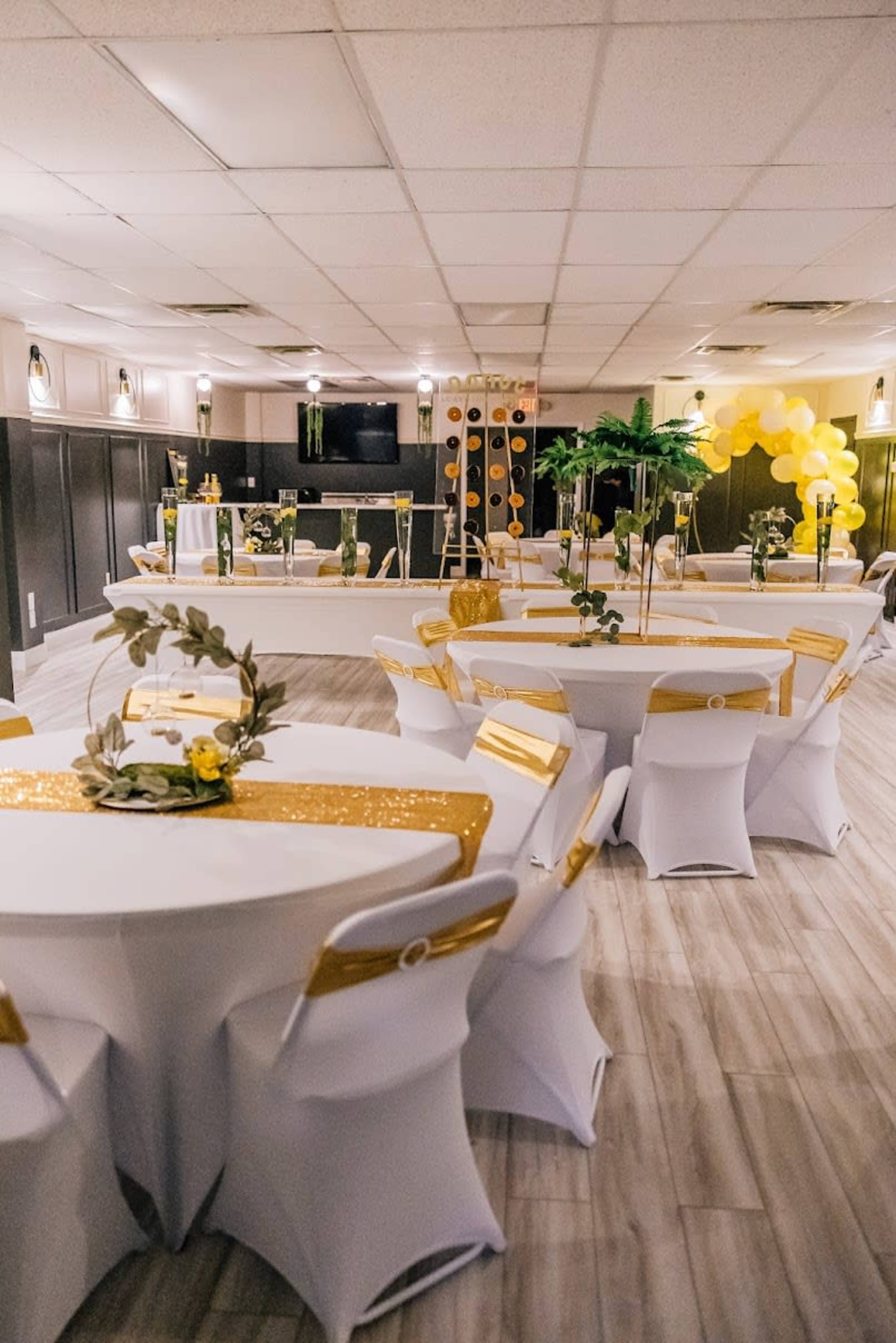A banquet hall is set up with round tables covered in white linens and gold accents, featuring decorations of greenery and yellow flowers.