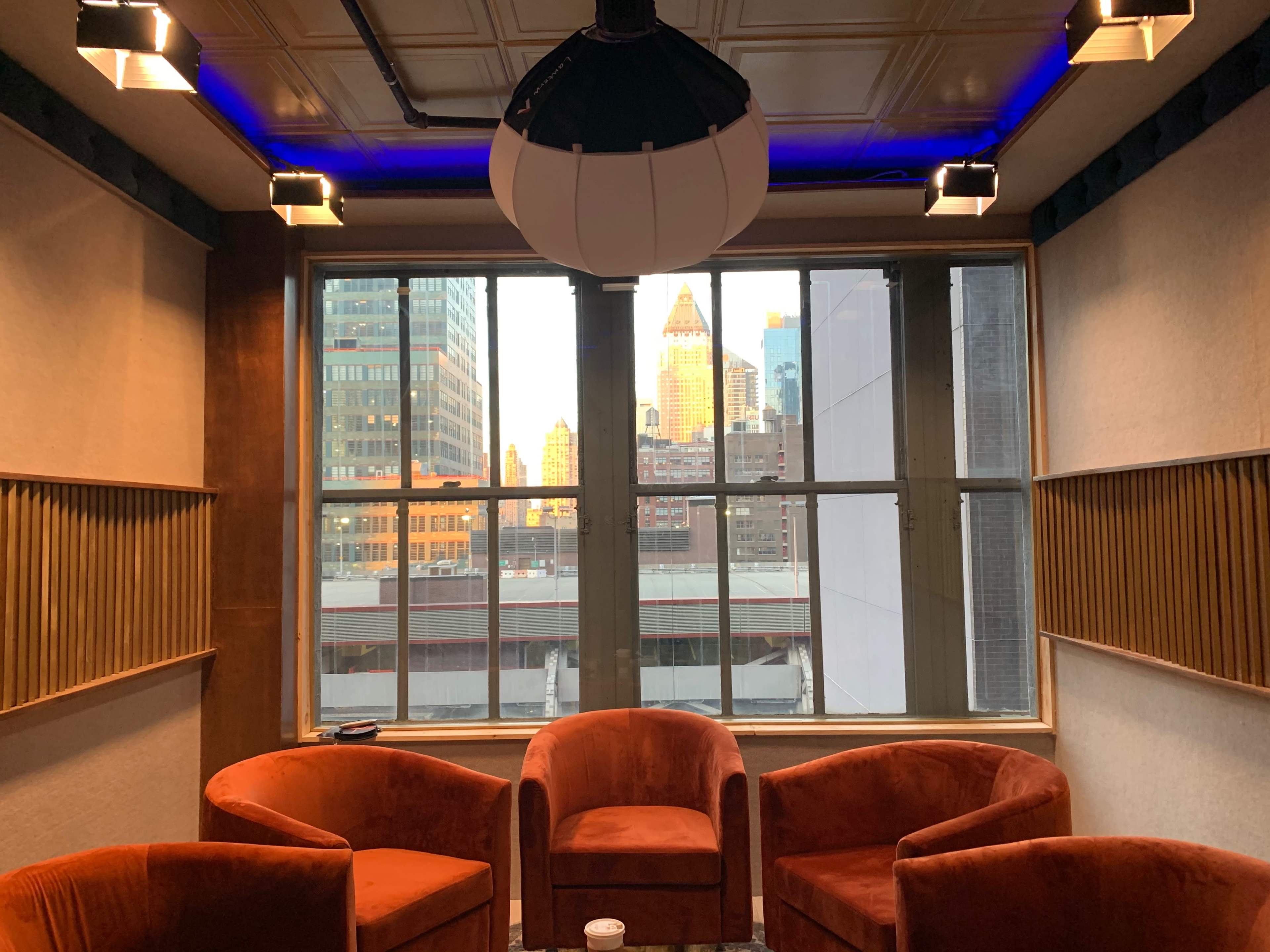 A cozy seating area with four orange chairs is positioned near a large window, providing a view of city skyscrapers at sunset.