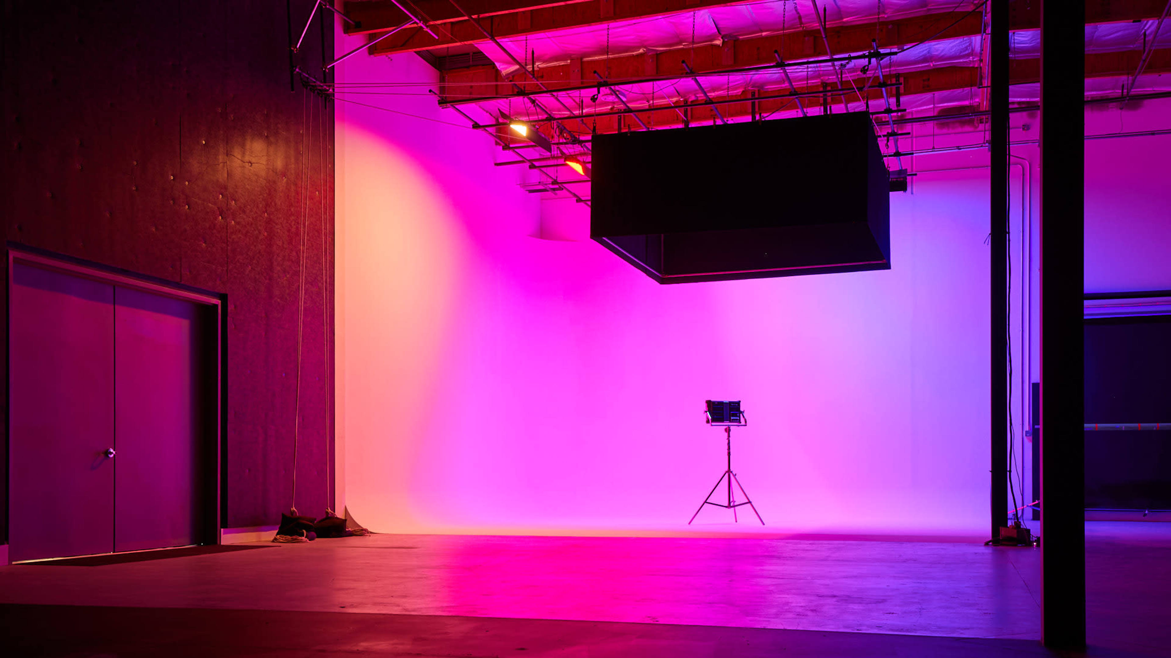 A spacious studio with a gradient backdrop of pink and purple lighting, featuring a light stand and a large black cube suspended above.