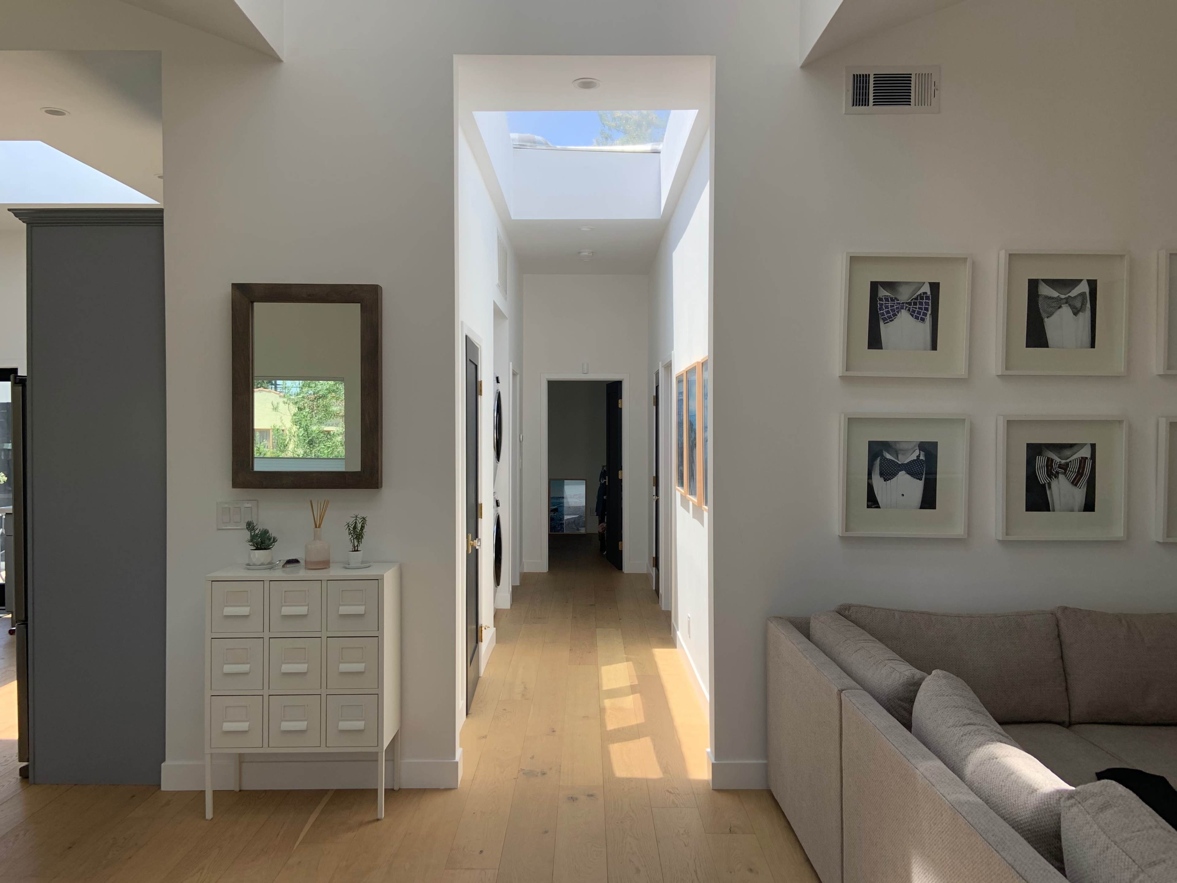 A bright hallway featuring a mirrored cabinet, framed artwork on the walls, and a view of an open interior space.