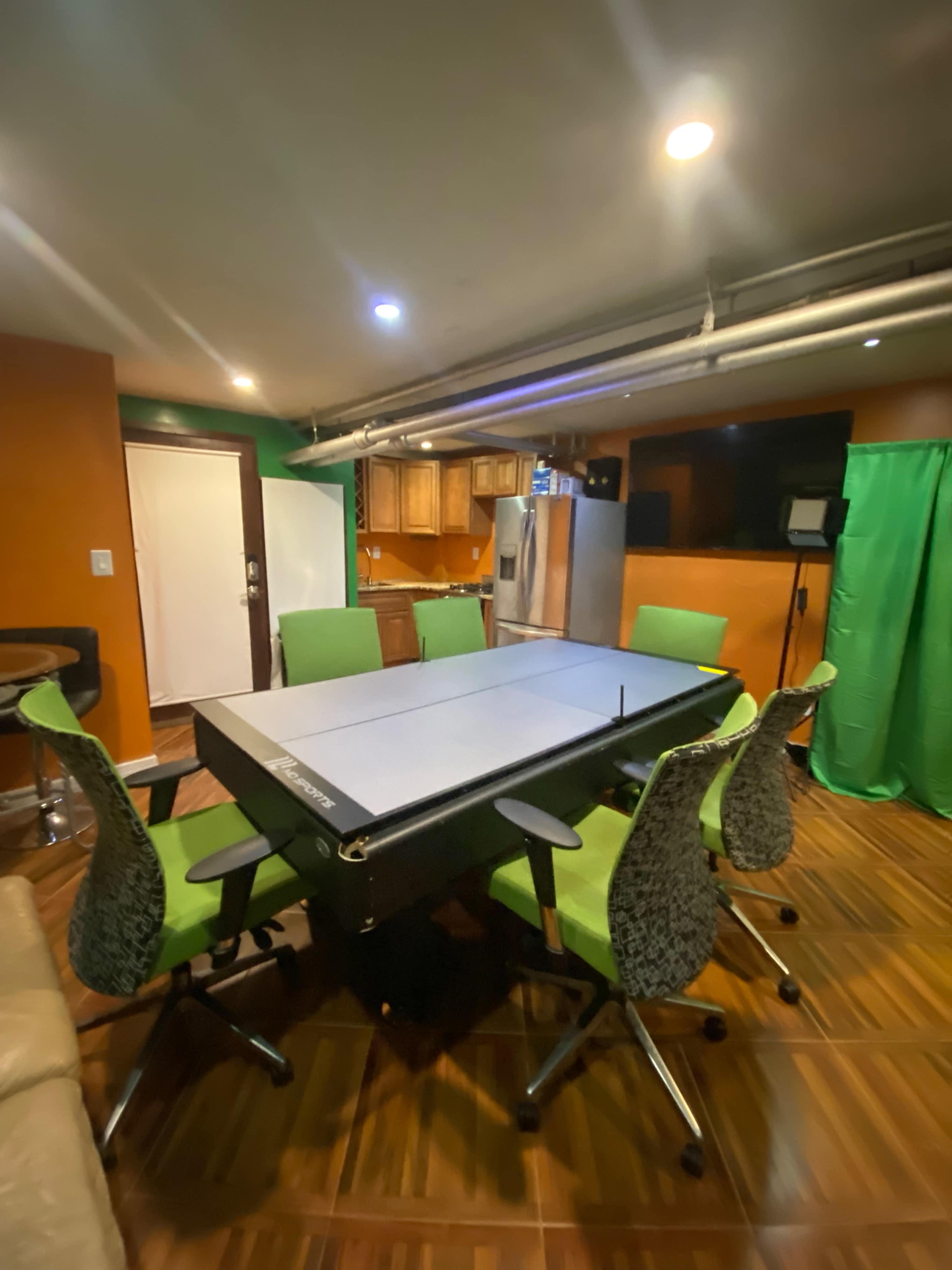 A table tennis table is surrounded by green office chairs in a room with orange walls and a kitchen area in the background.