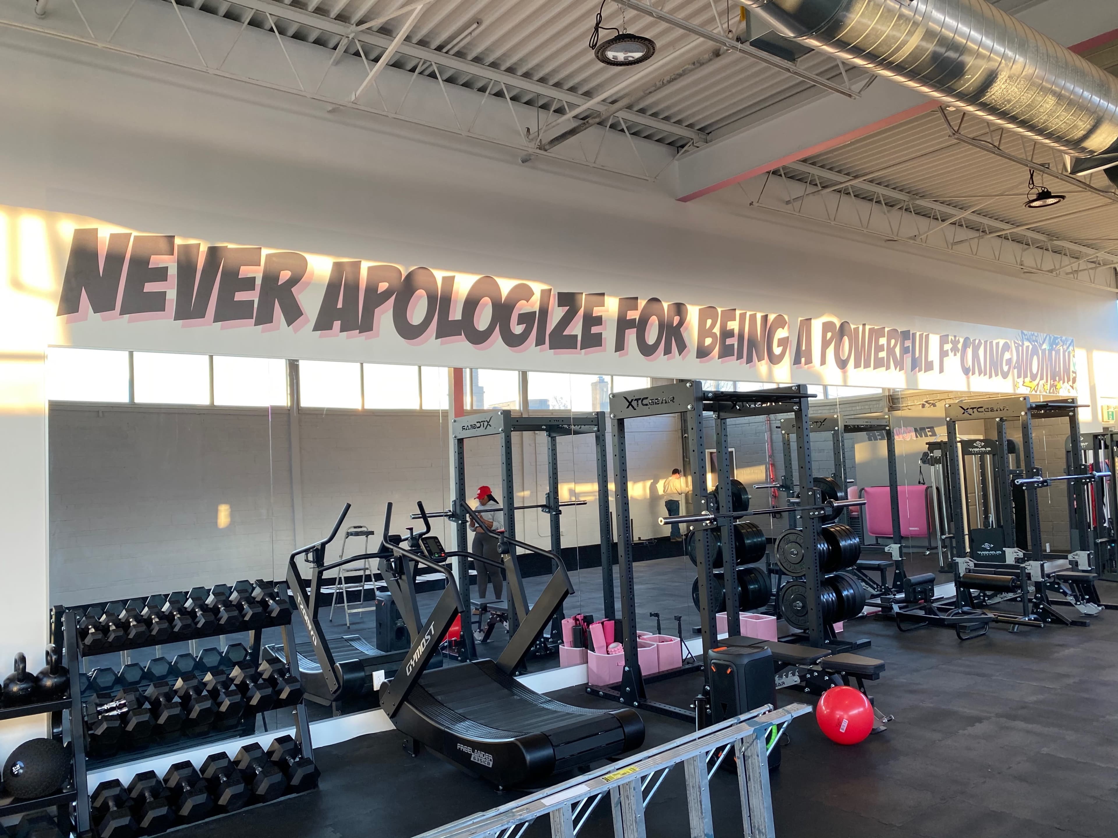 The interior of a gym features weightlifting equipment and a motivational phrase displayed on the wall.