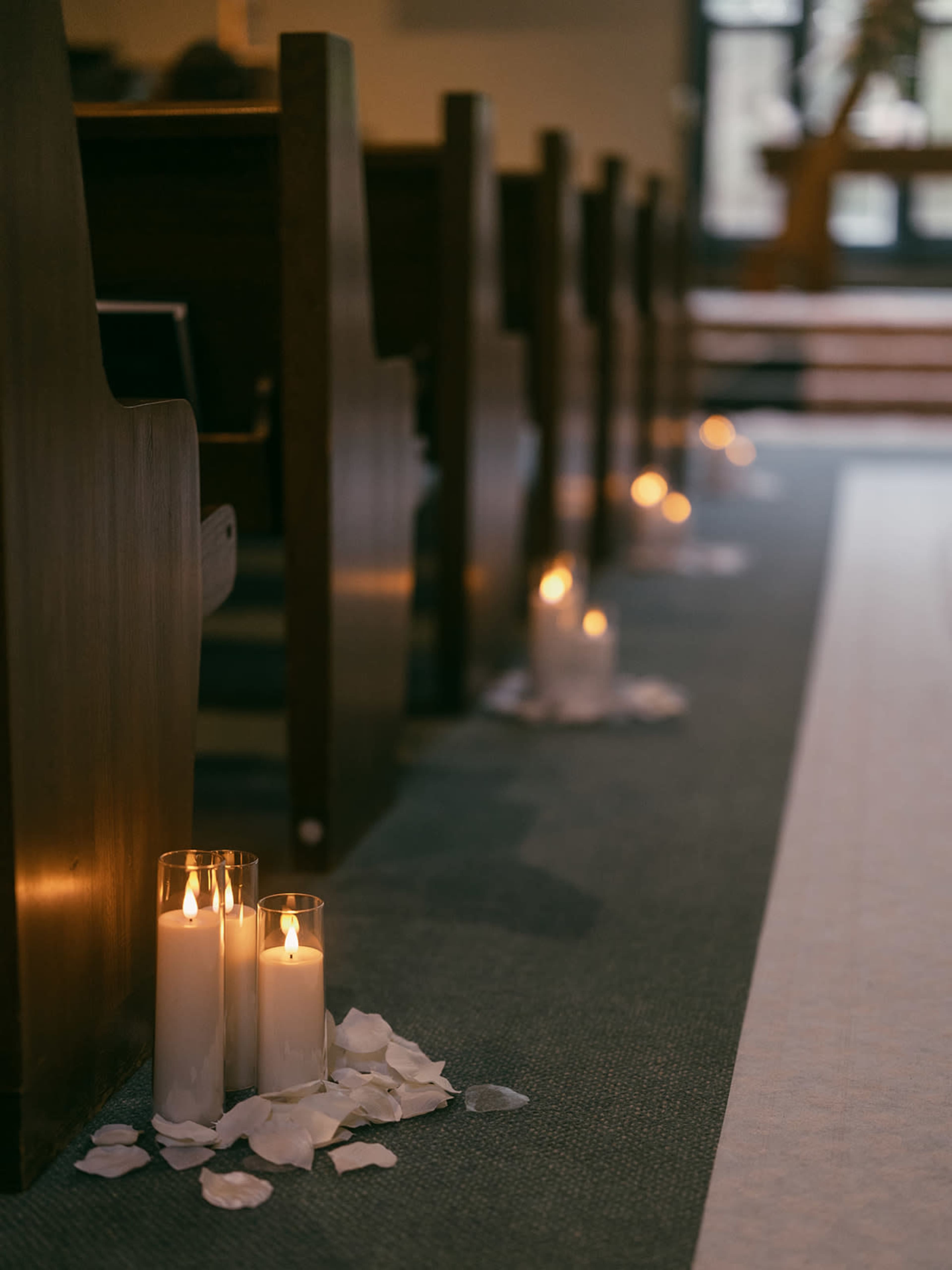 A row of church pews with lit candles and rose petals arranged along the aisle.