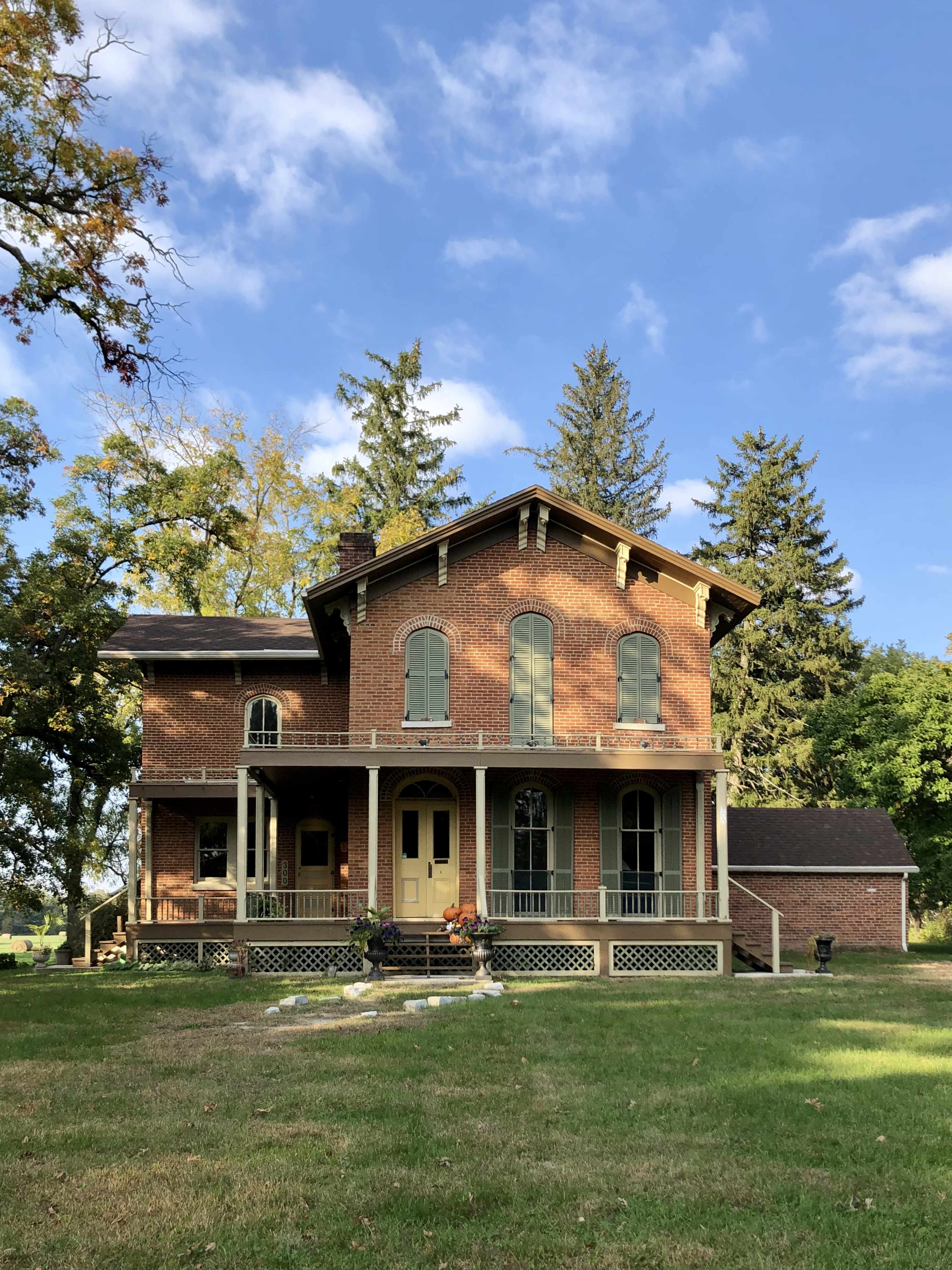 A two-story brick house with large windows and a front porch is surrounded by tall trees and a grassy yard.