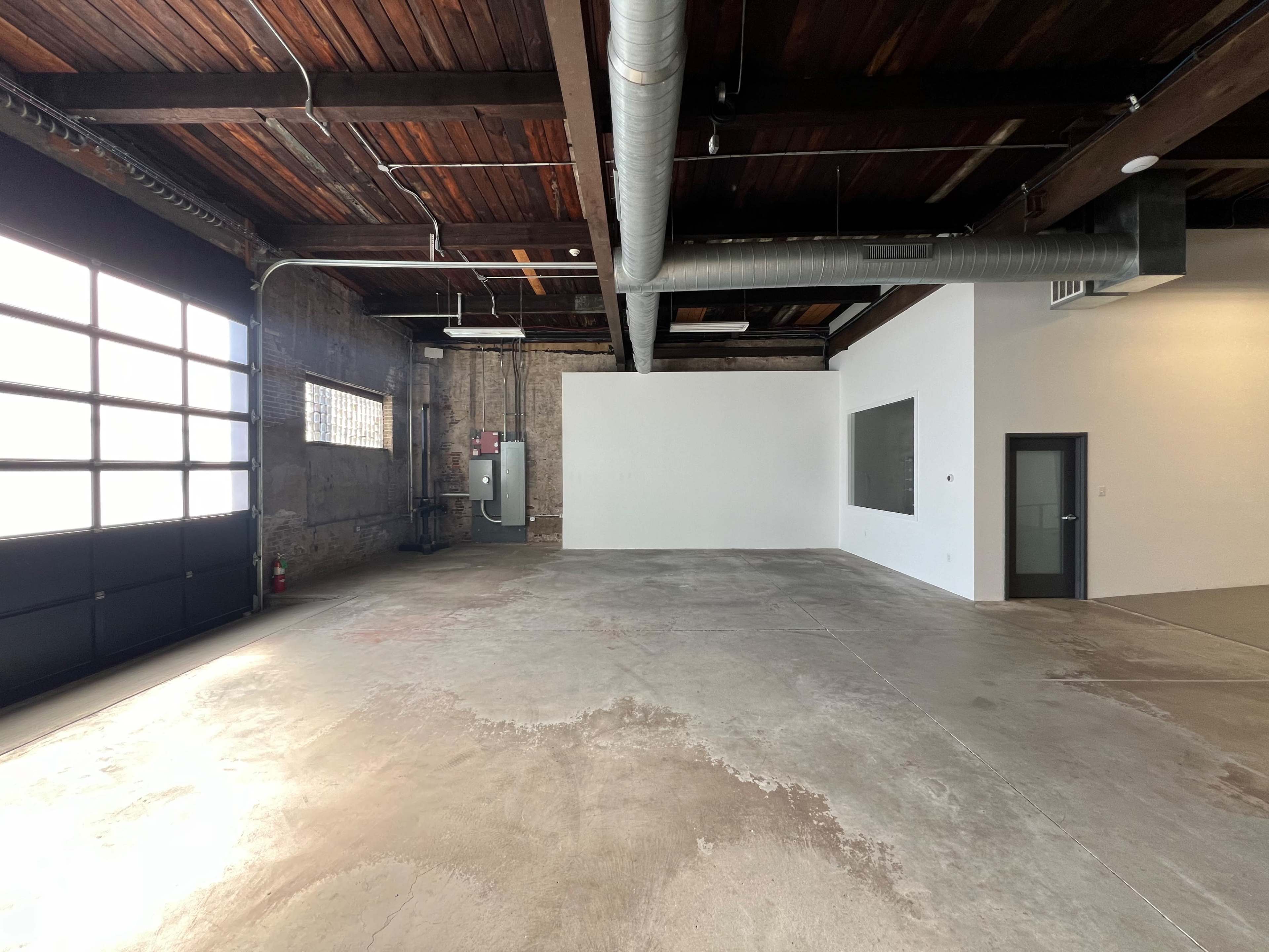 A spacious, empty industrial-style room with exposed wooden beams, a concrete floor, and large windows.