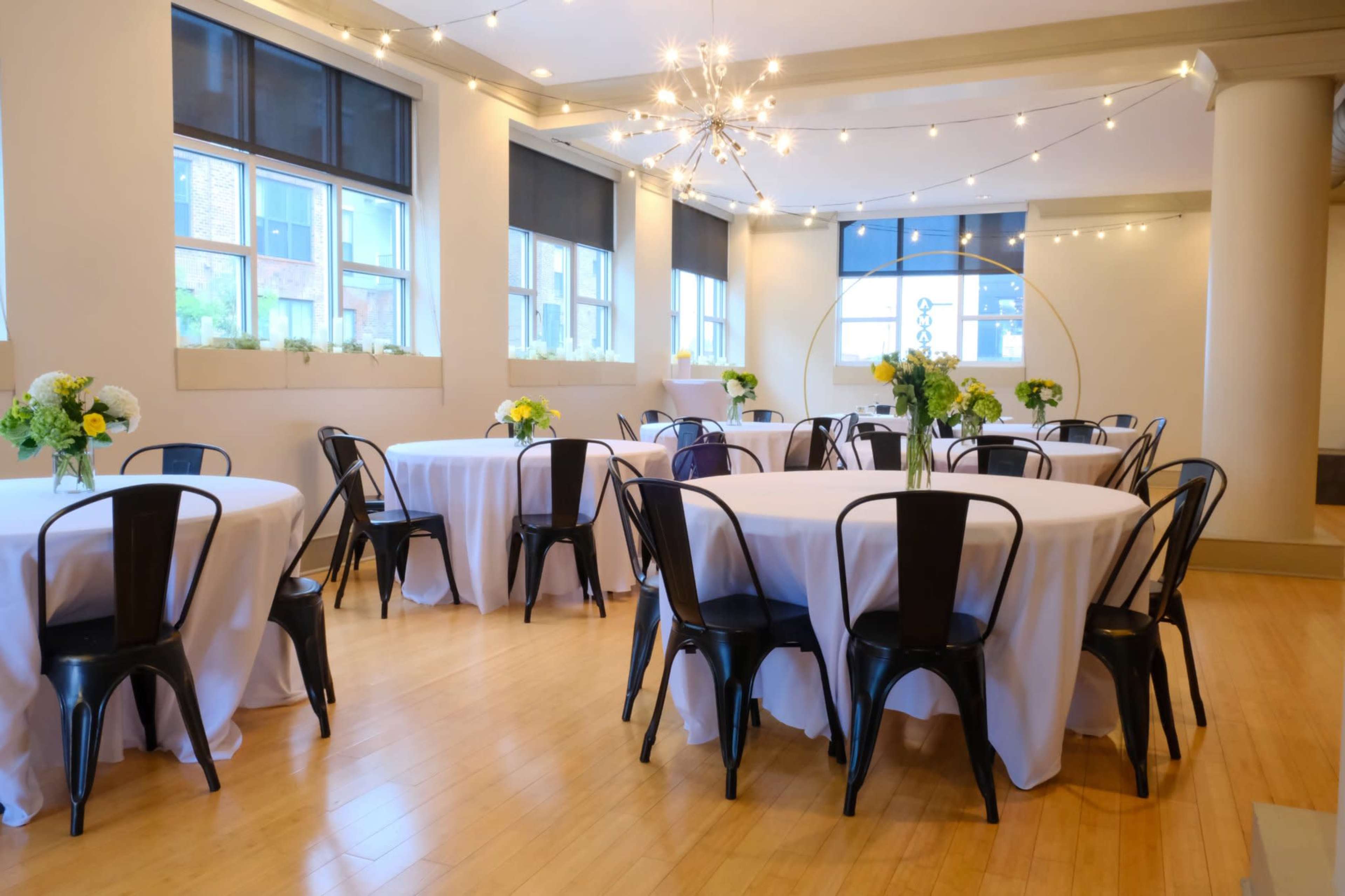 A elegantly arranged event space with round tables covered in white tablecloths and black chairs, illuminated by string lights overhead.