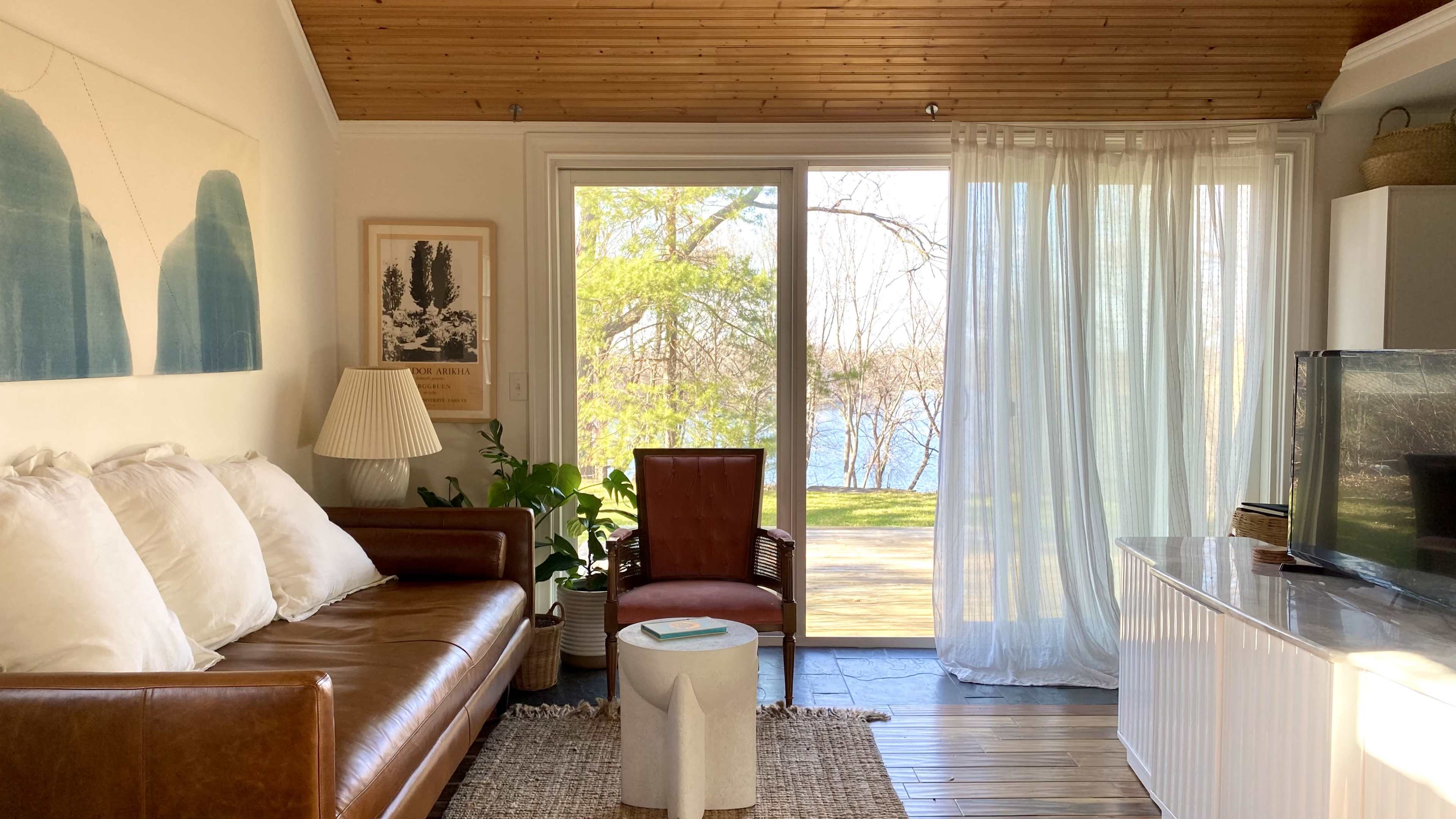A cozy living room features a brown leather sofa, a wooden chair, a round white table, and large windows with sheer curtains overlooking a lake.