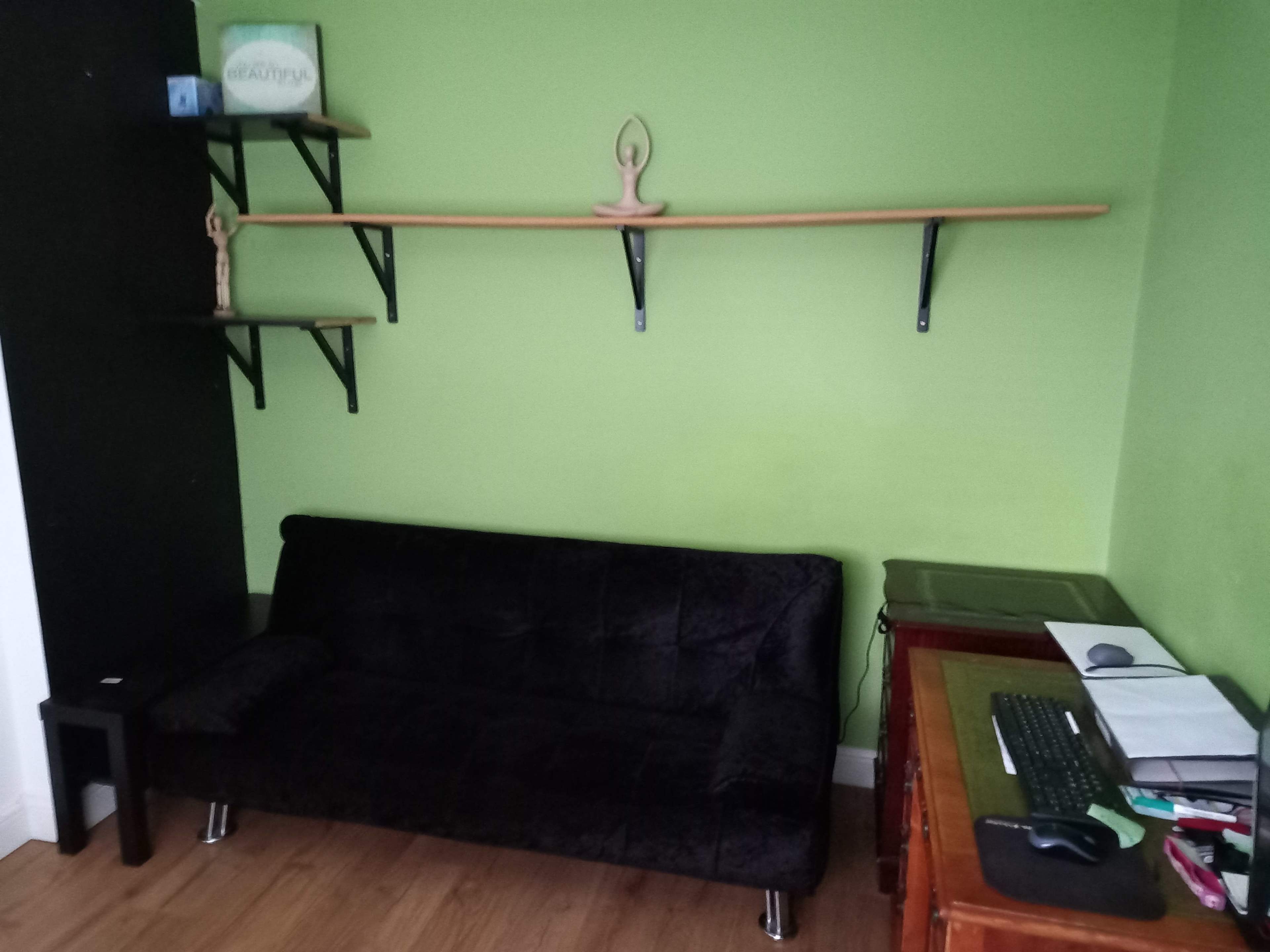 A black sofa against a green wall, with a wooden shelf above it and a desk with a keyboard and papers to the right.