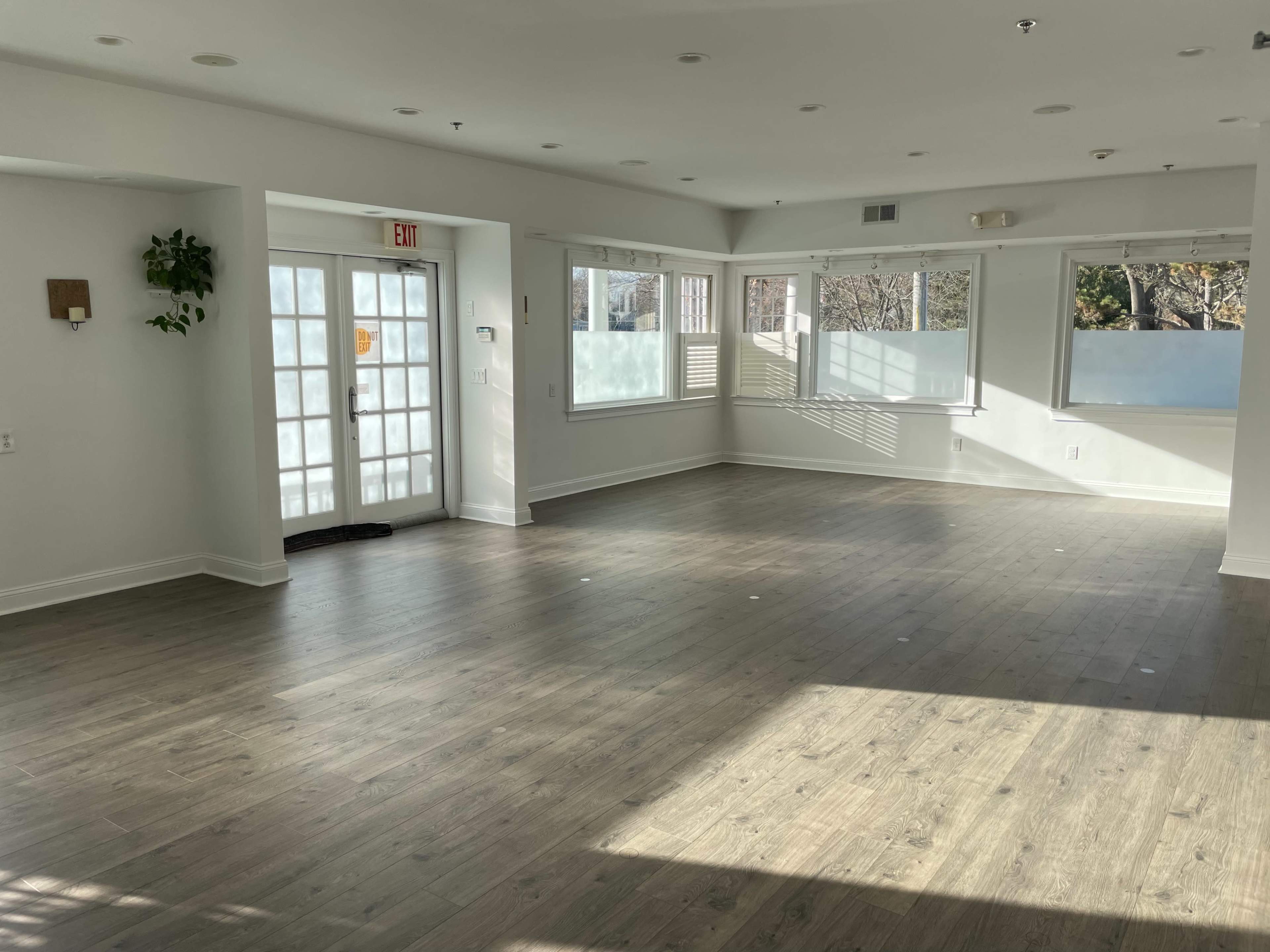 A spacious, well-lit room with wooden flooring, large windows, and a glass door leading outside.