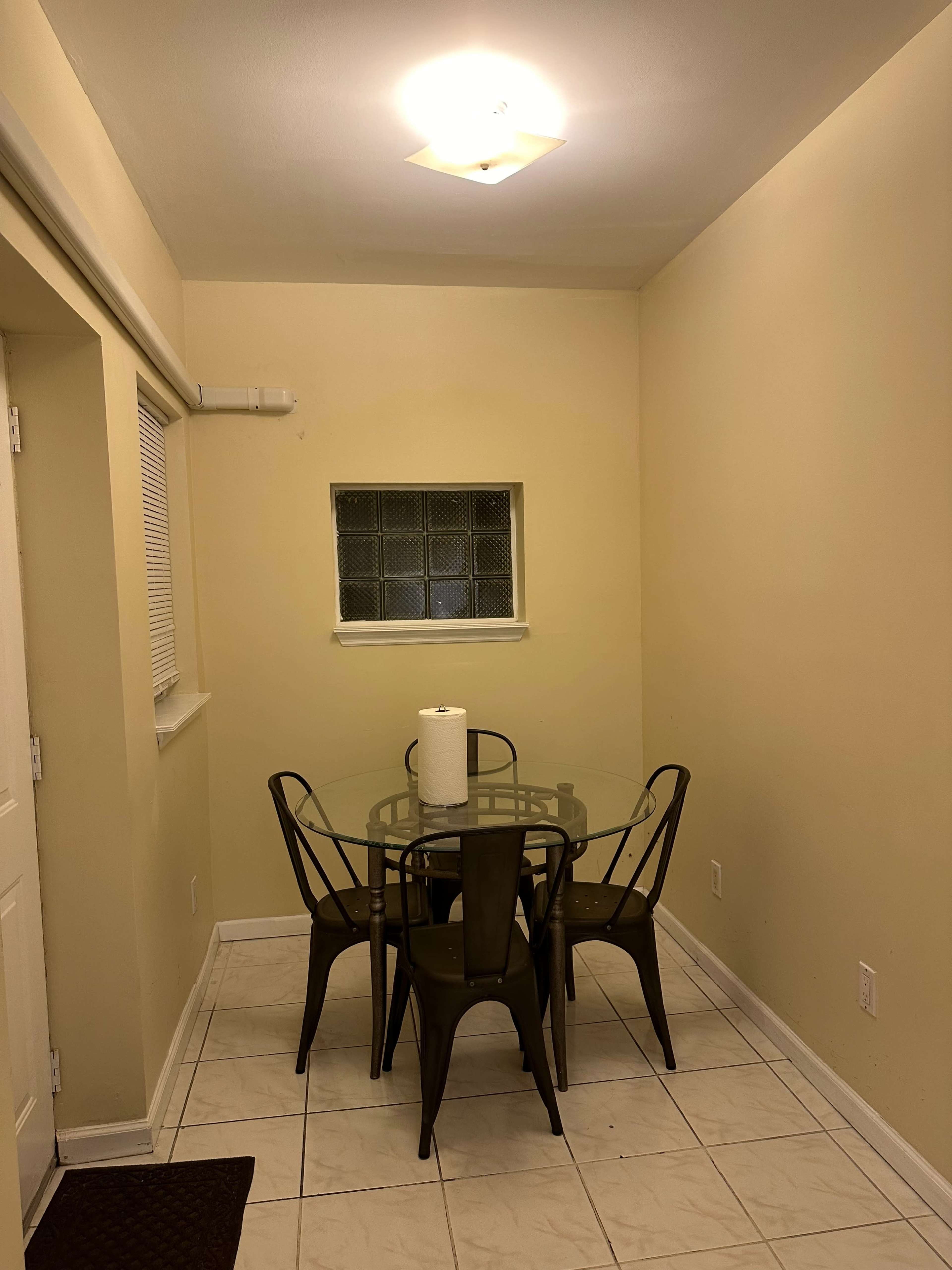 The image shows a small dining area with a glass table, four metal chairs, and a paper towel roll on the table, set against light-colored walls and a window.