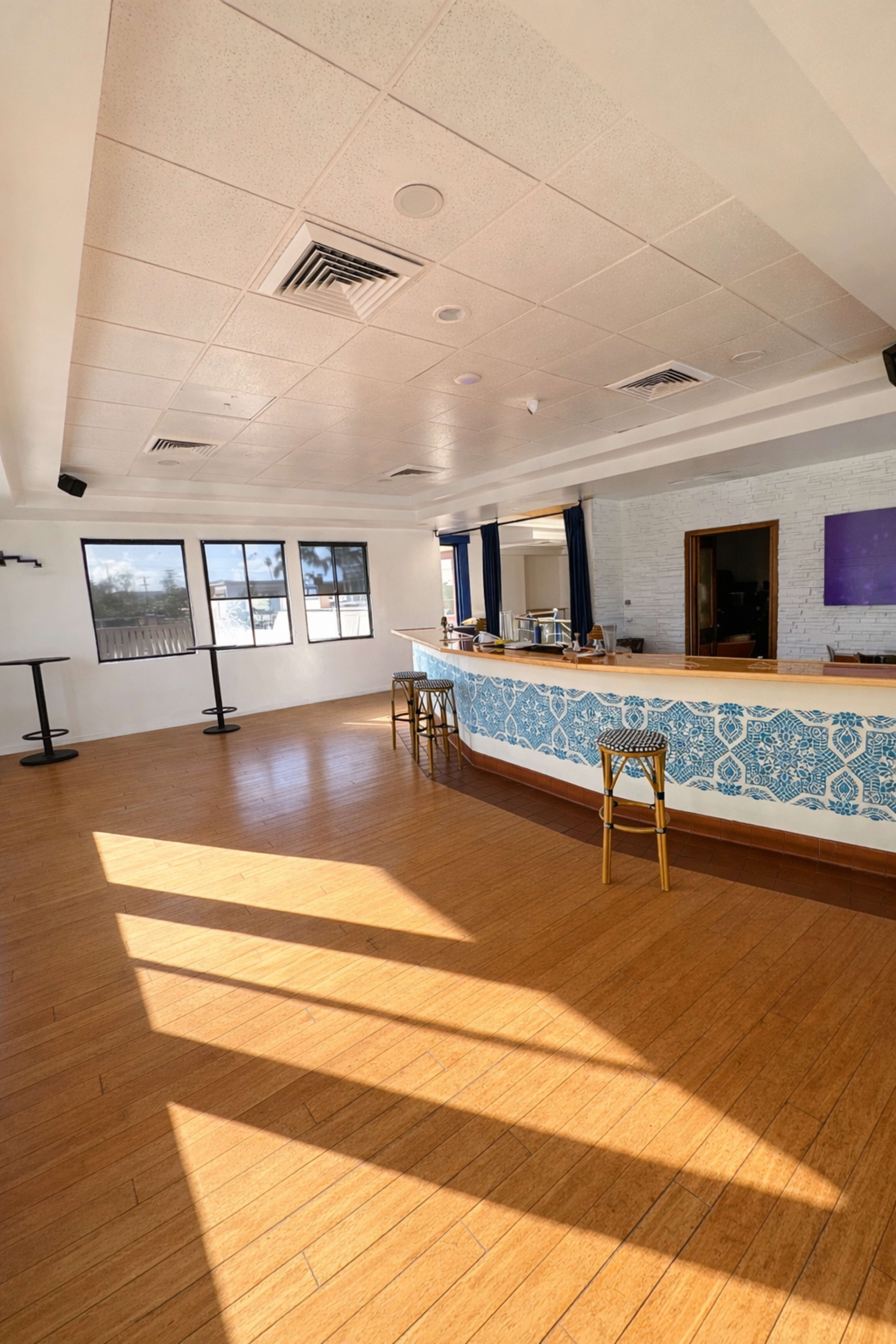 The image shows a spacious, brightly lit interior of a bar featuring a curved blue tile counter and wooden flooring, with shadows cast across the room.