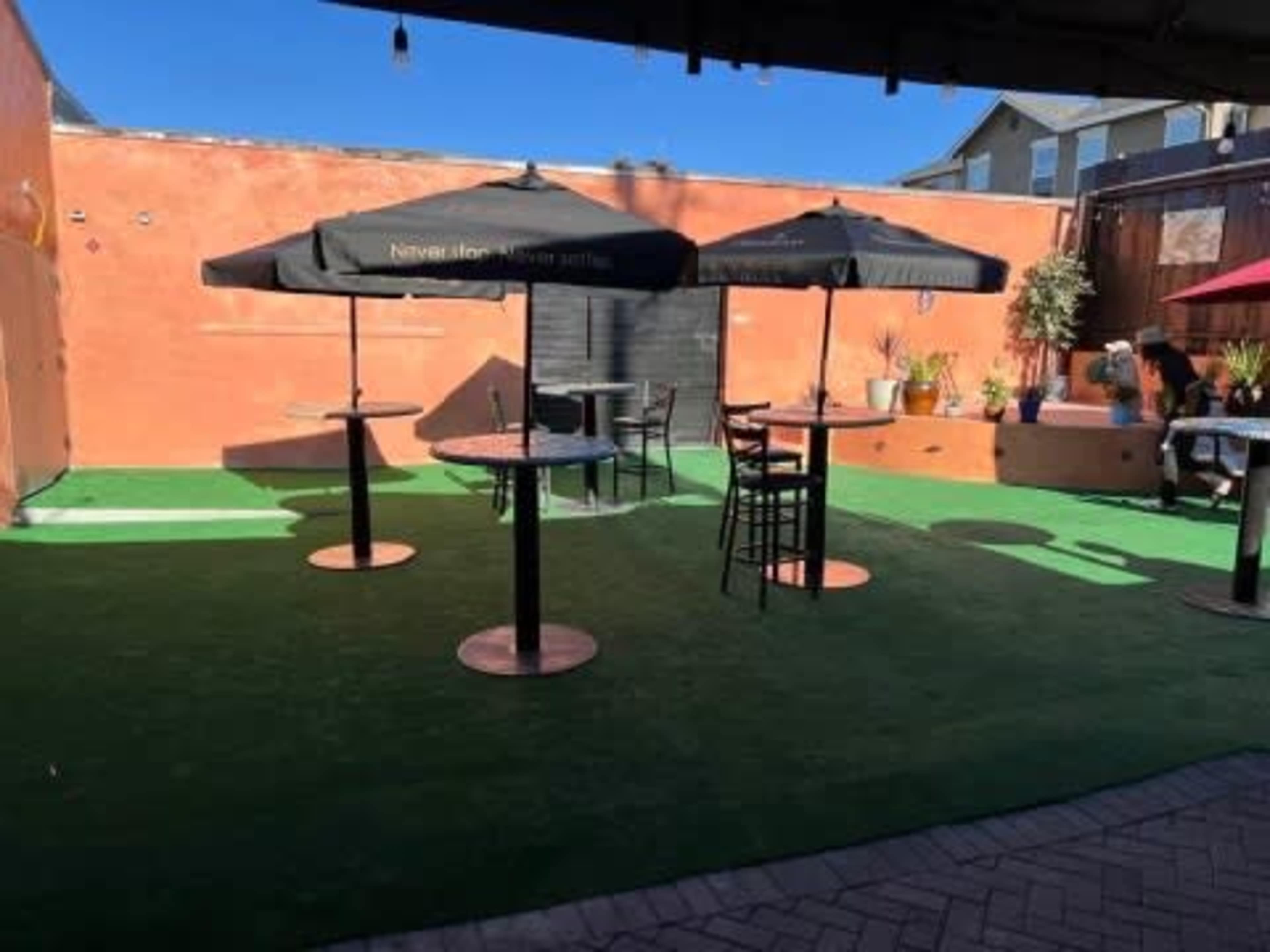 The image shows an outdoor area with green turf, several black umbrellas over tables, and a few potted plants against a colorful wall.