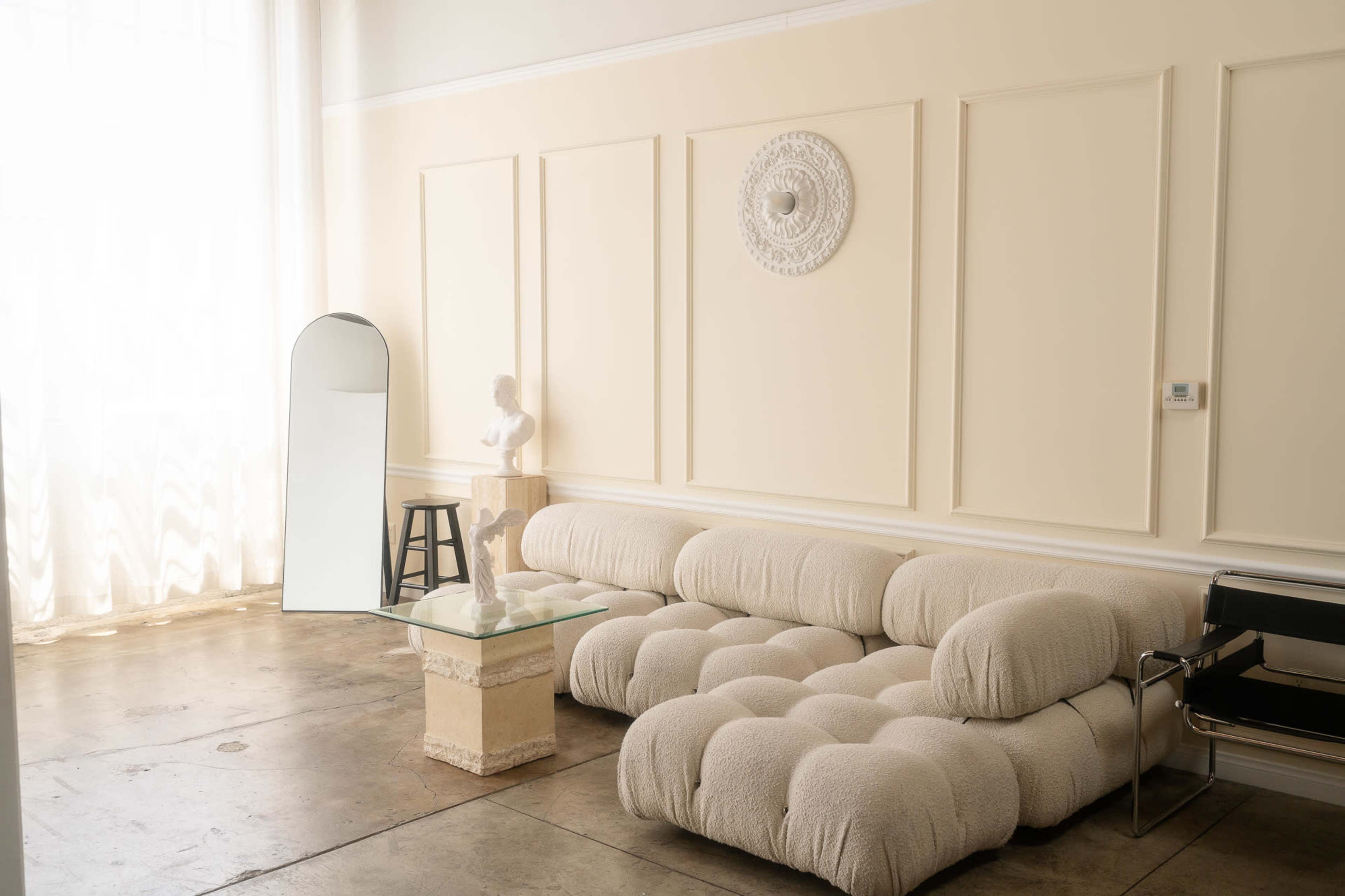 A light-colored living room with a textured sectional sofa, a glass coffee table, a mirror, and a sculpture displayed on a pedestal.