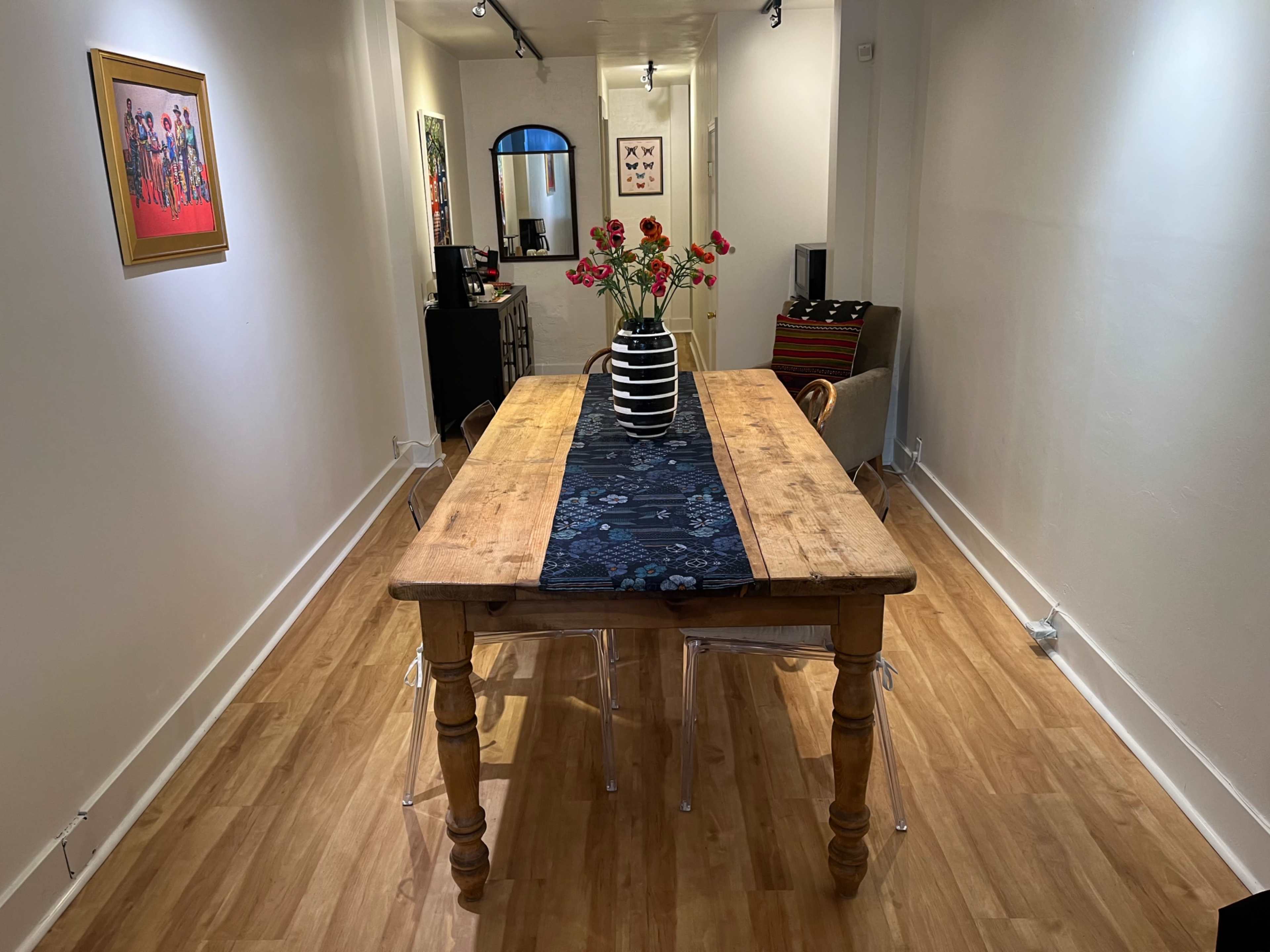 A wooden dining table is set in a corridor with a vase of flowers in the center, flanked by a chair and various artworks on the walls.