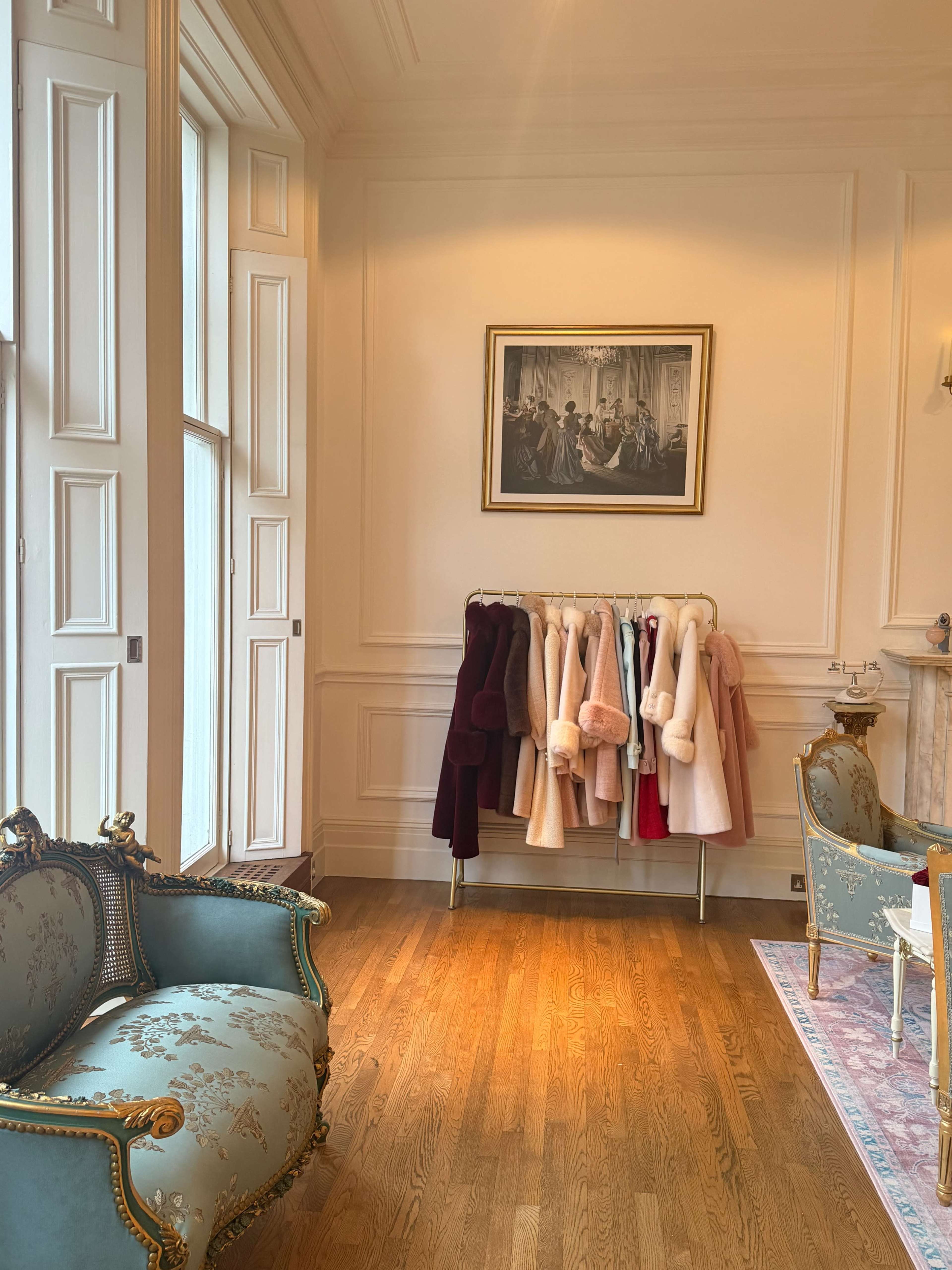 A neatly arranged display of coats hangs on a rack against a light-colored wall in a well-furnished room featuring ornate furniture and a framed painting.