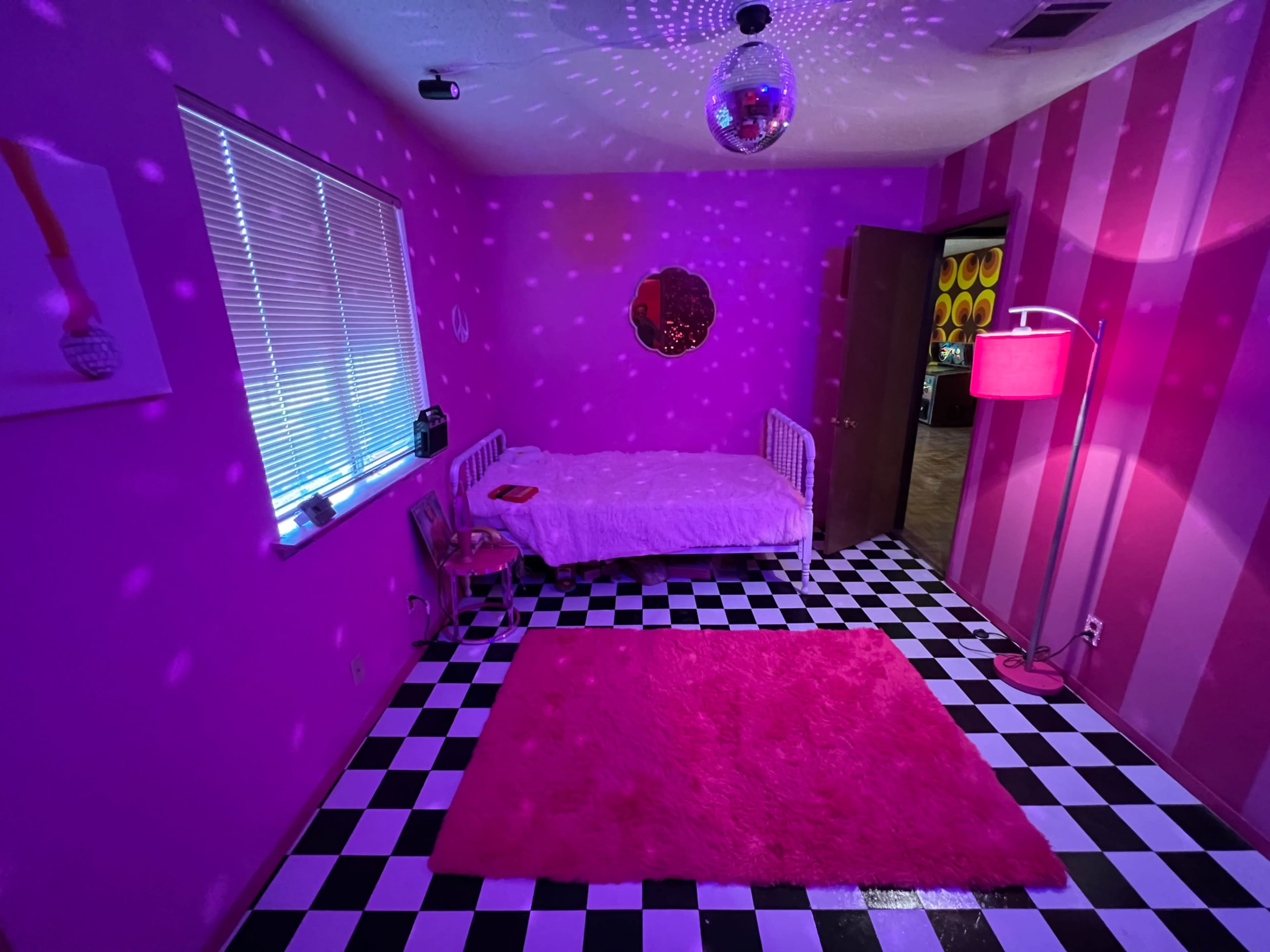 The image depicts a vibrant pink room with a disco ball, striped walls, a white bed, and a black-and-white checkered floor.