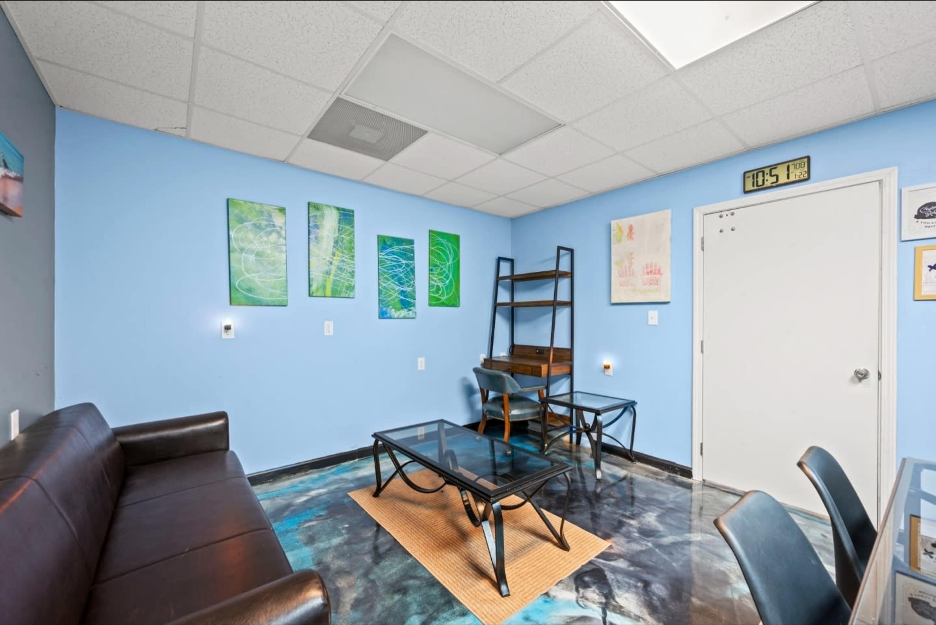 The image shows a small room with blue walls, a brown couch, a glass coffee table, and artwork hanging on the walls.