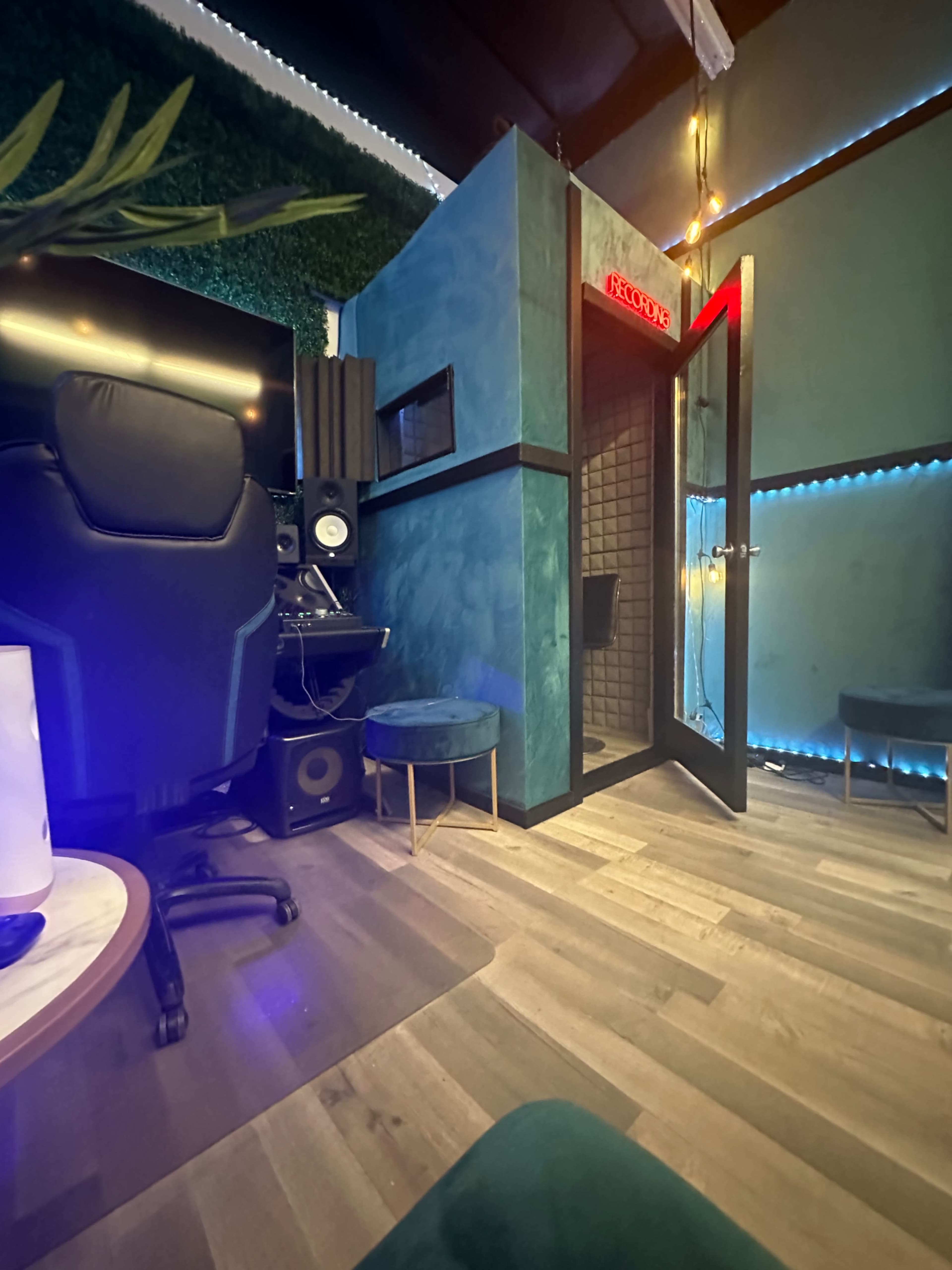 A recording studio interior features a soundproof booth, a computer setup with audio equipment, and modern seating.