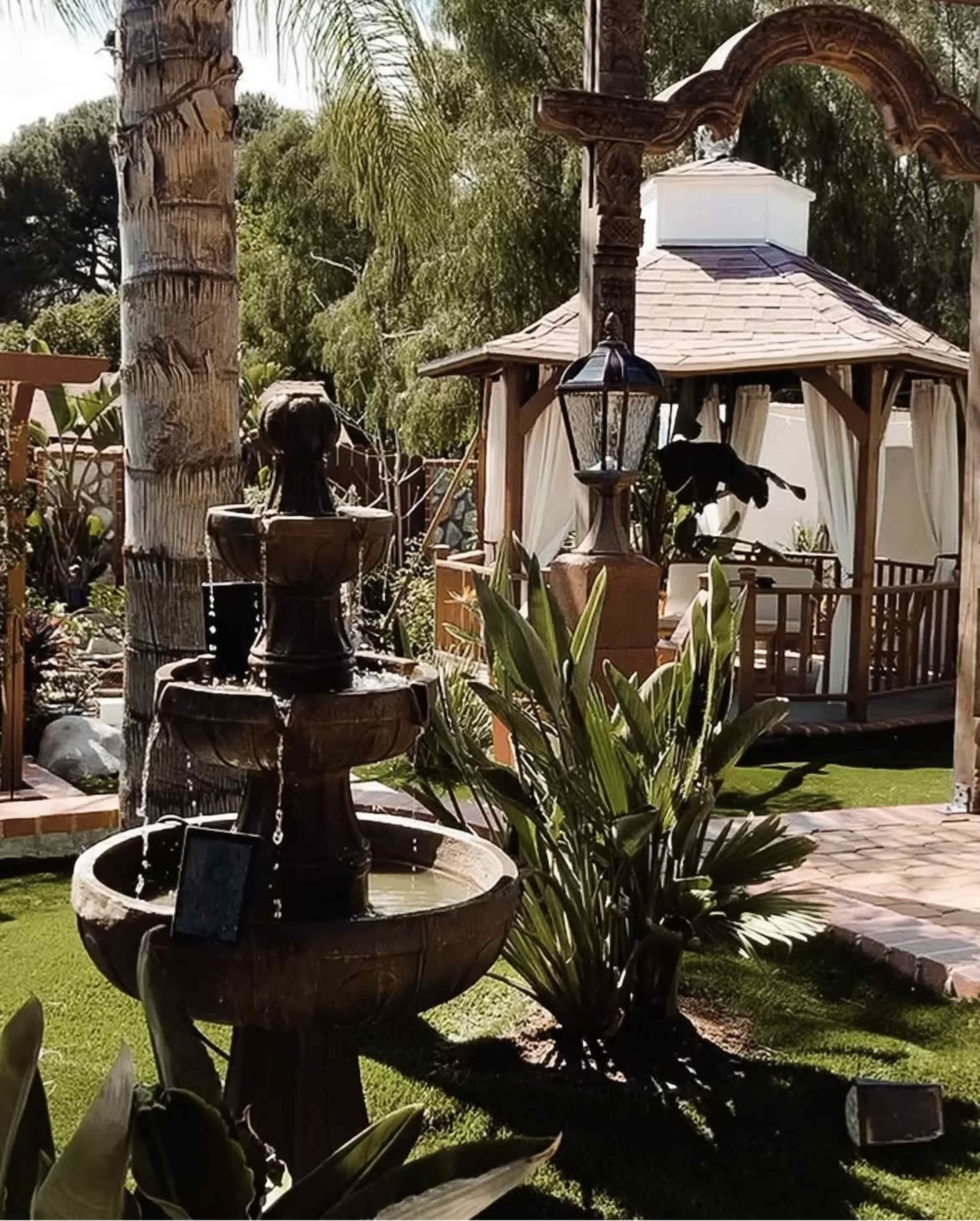 The image shows a tiered fountain surrounded by lush greenery and a gazebo in a well-maintained garden.