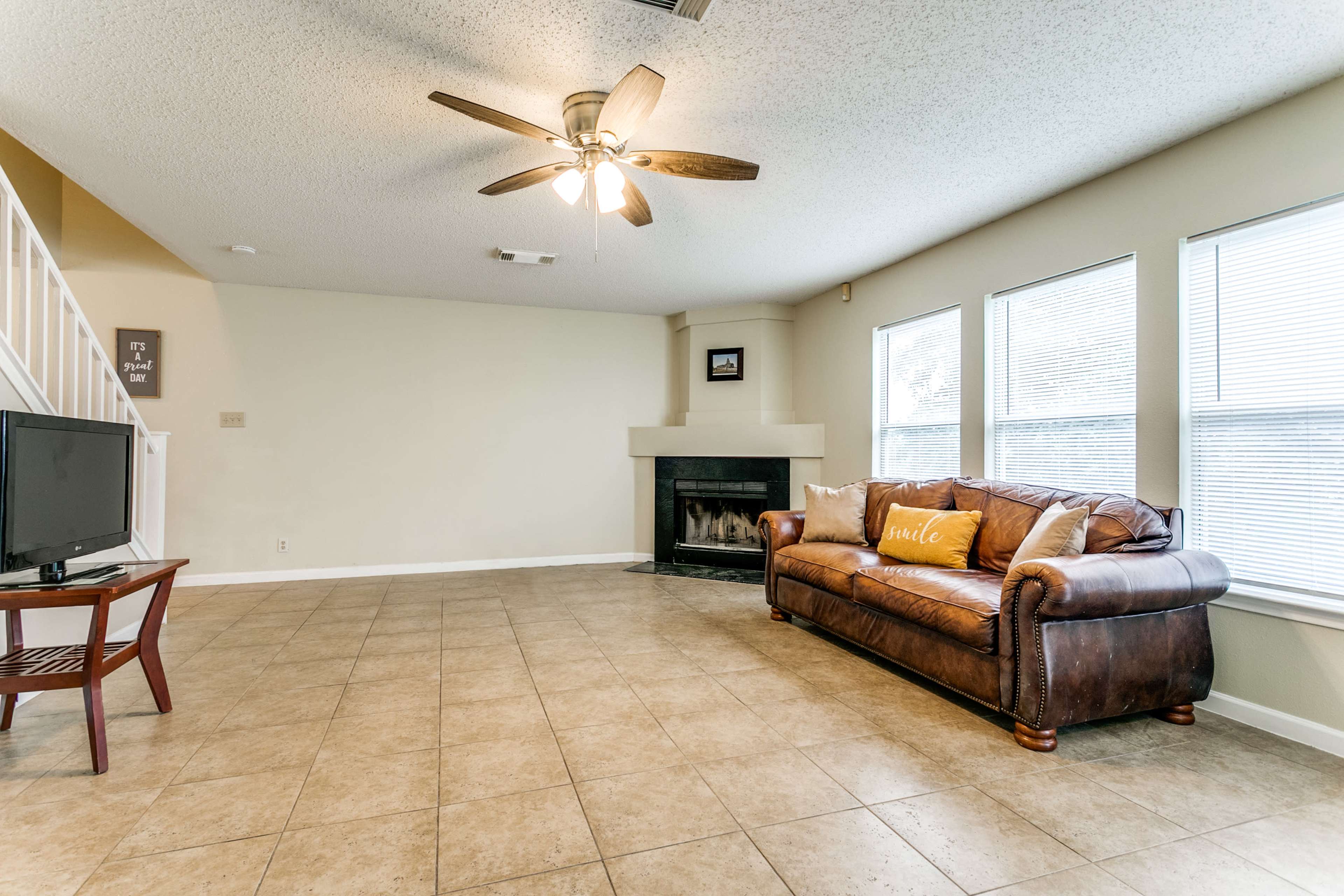 A spacious living room with tile flooring, a brown leather sofa, a television on a stand, and a fireplace under a large window.