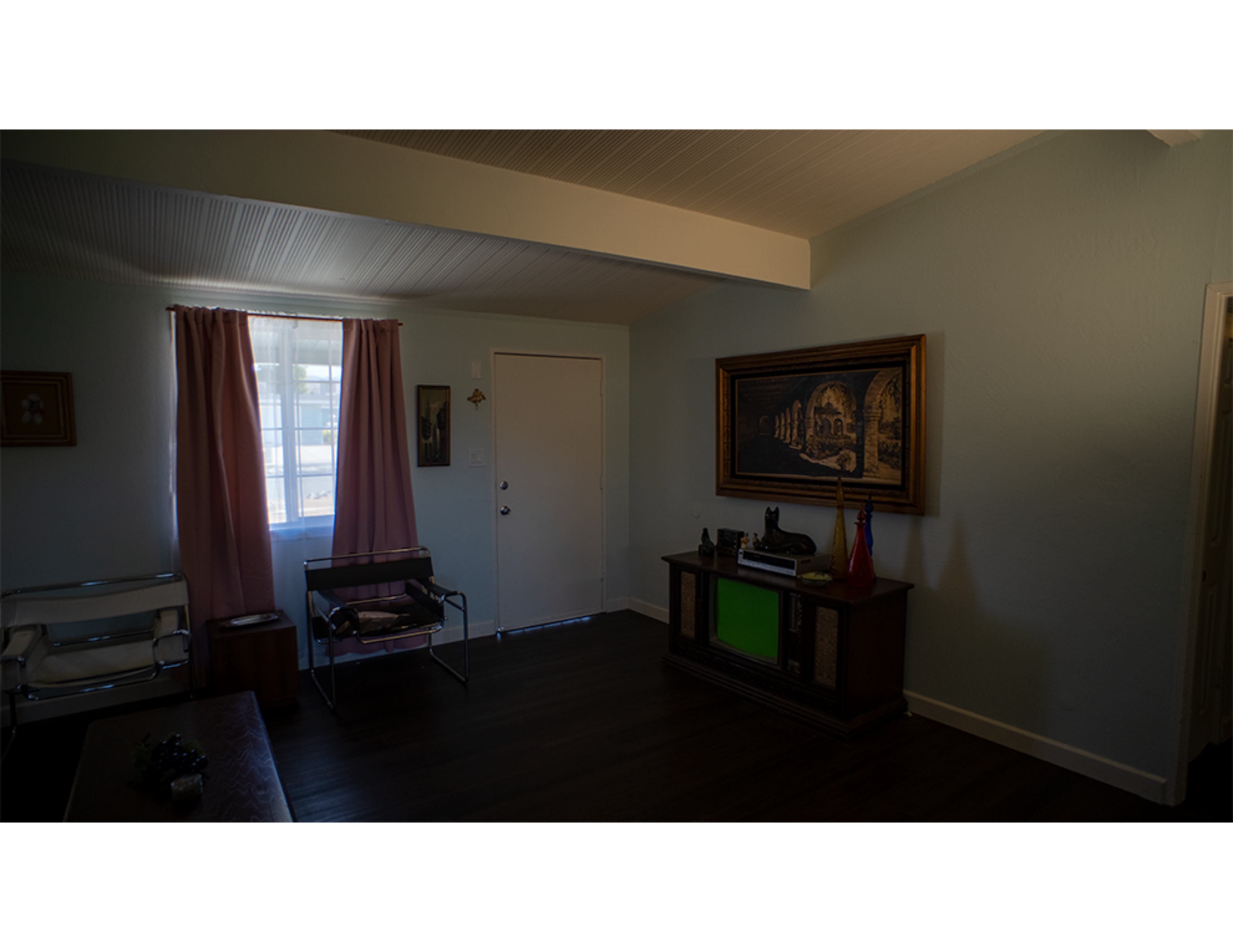 A sparsely furnished living room with a door and window, framed by soft curtains, and a large picture hanging on the wall.